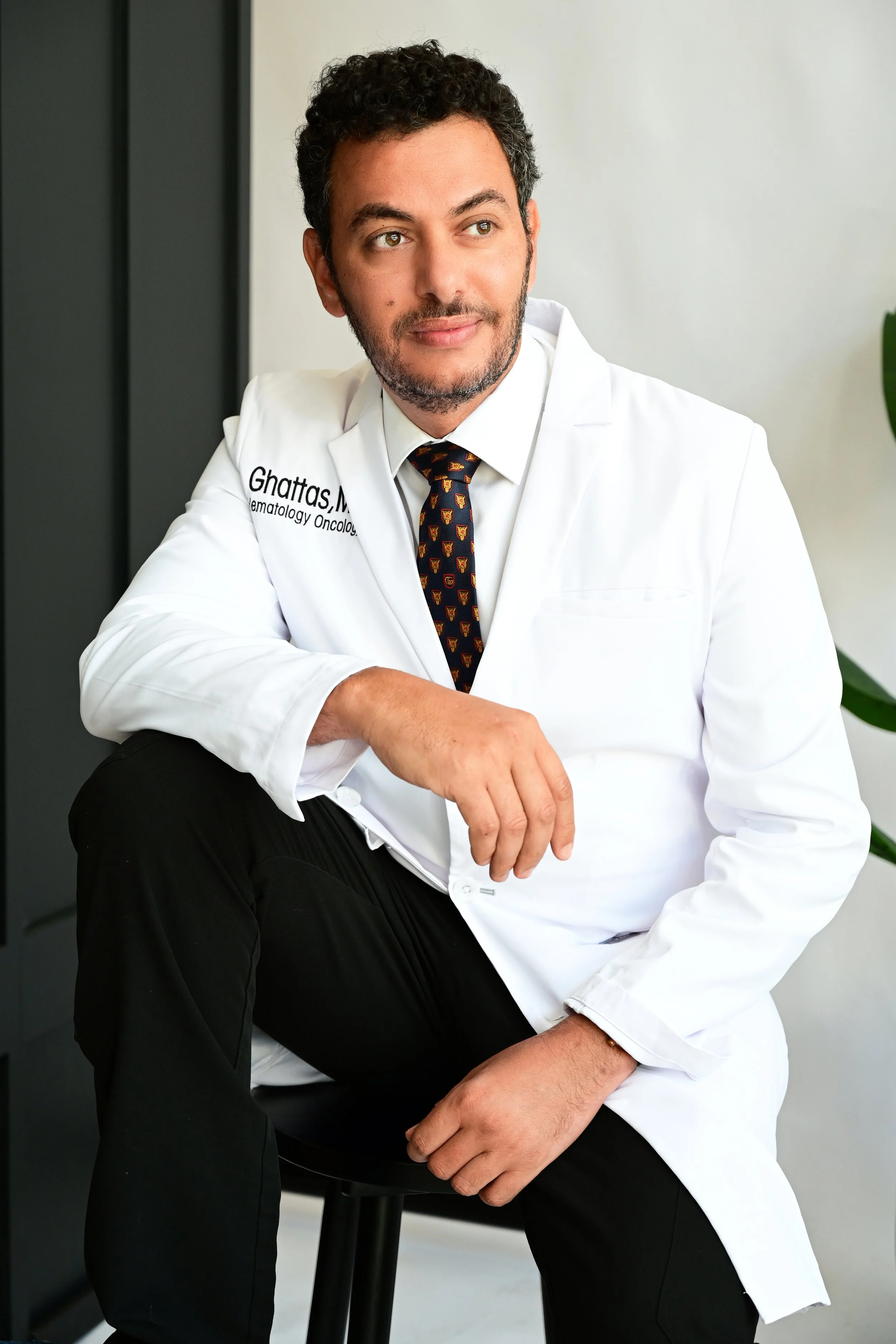 A man with curly hair and a beard sitting on a stool in a professional setting. He is wearing a white coat with text on it, a shirt, and a patterned tie. He appears to be a medical professional, possibly a doctor.