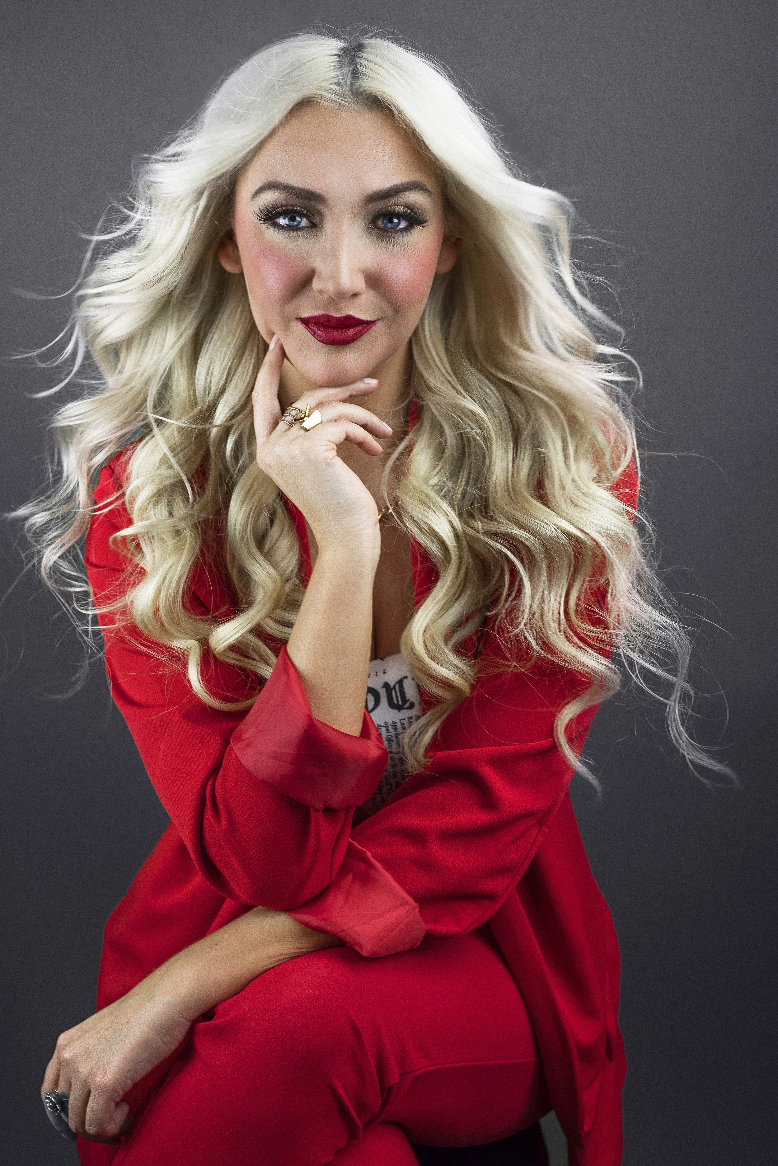 A woman with long, blonde, wavy hair and blue eyes posing with her hand near her chin on a gray background. She is wearing a red jacket and matching red pants, with bold makeup including dark eyeliner, long lashes, and red lipstick.