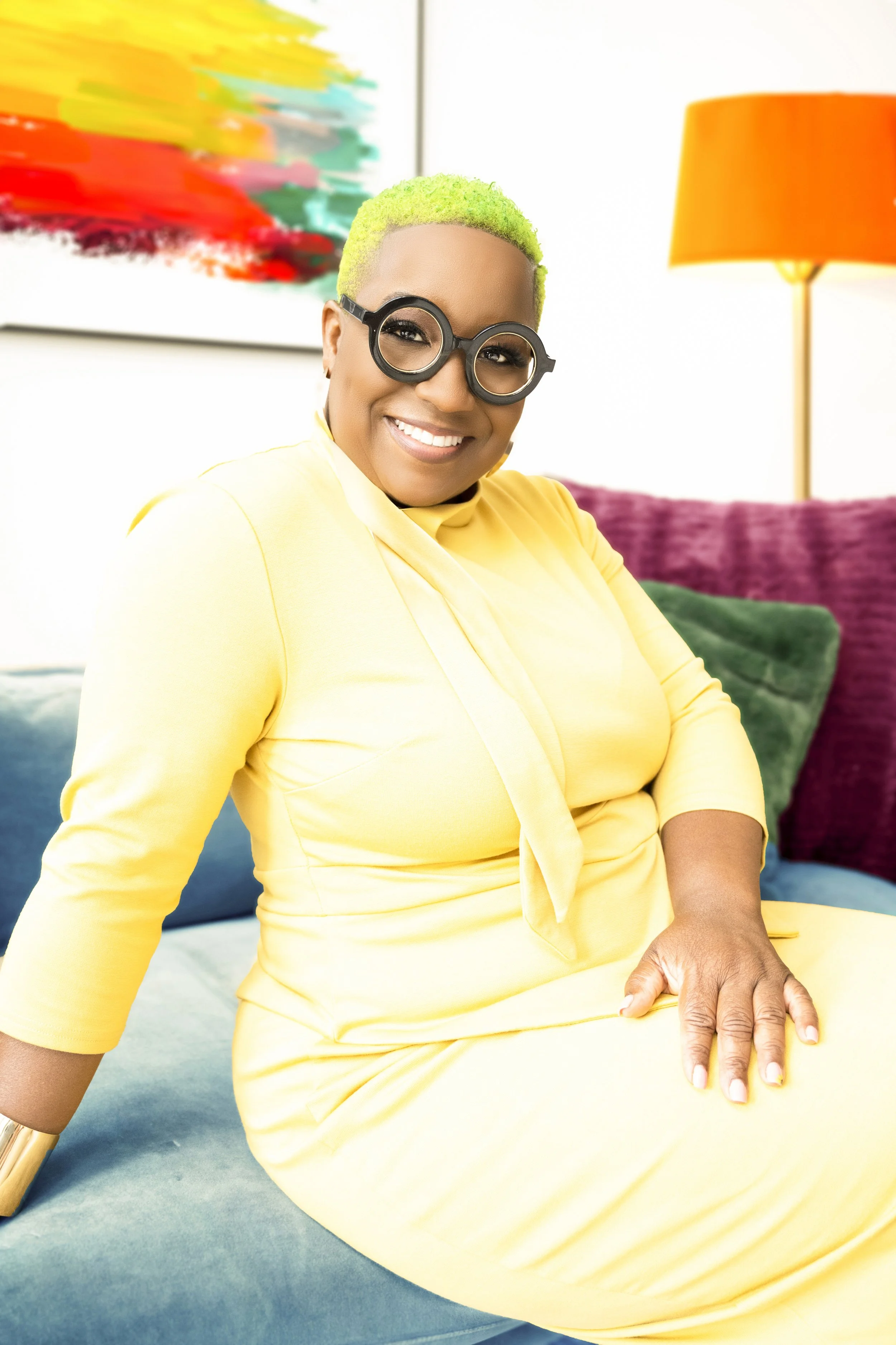 Woman with short, bright green hair wearing large, round black glasses, yellow dress, sitting on a teal couch in a brightly lit room, with colorful artwork and a yellow lamp behind her.