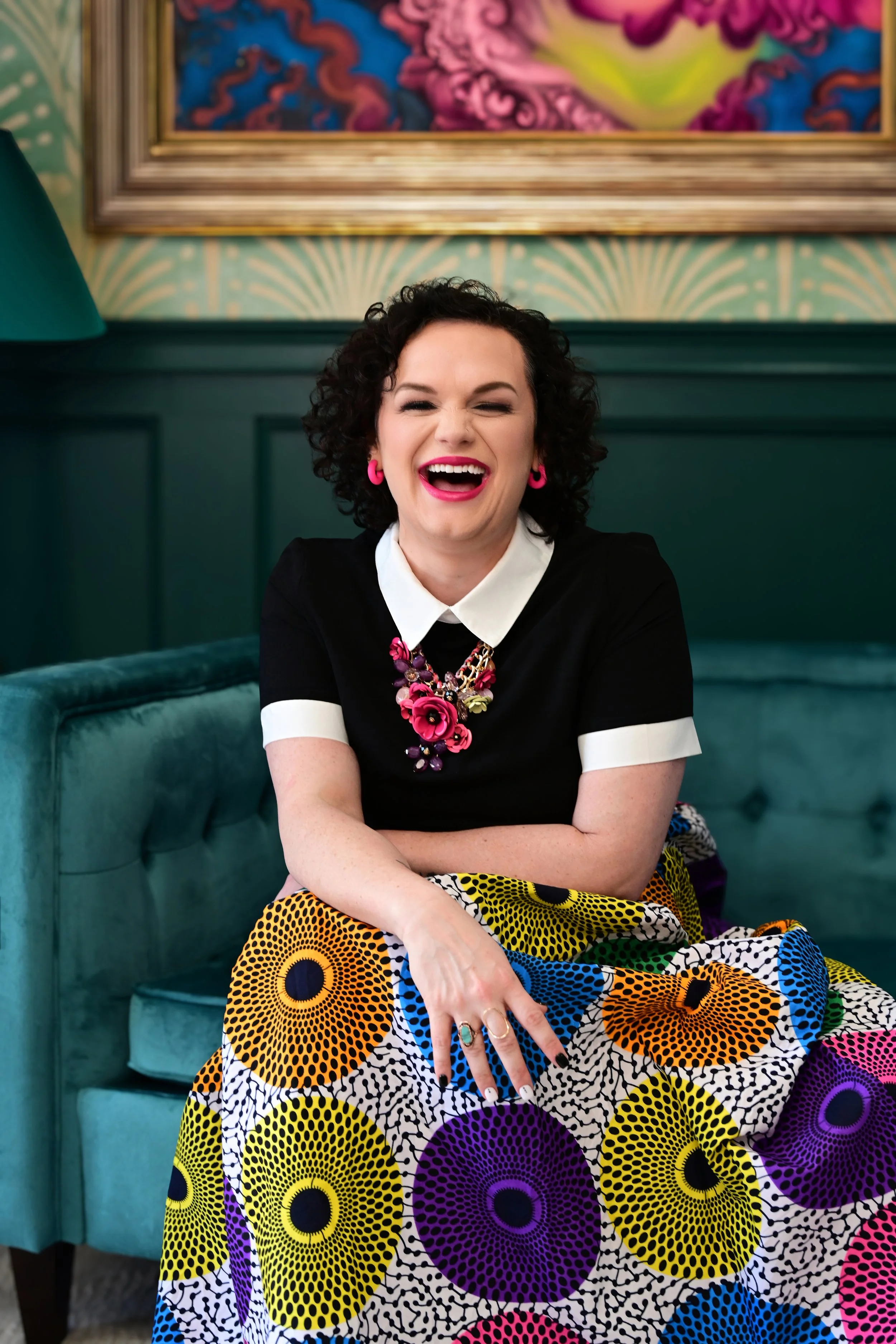A woman with short curly black hair, wearing pink earrings and a floral necklace, sitting on a teal velvet sofa. She is laughing with her eyes closed, wearing a black dress with white collar and sleeve cuffs, and a colorful patterned skirt with large