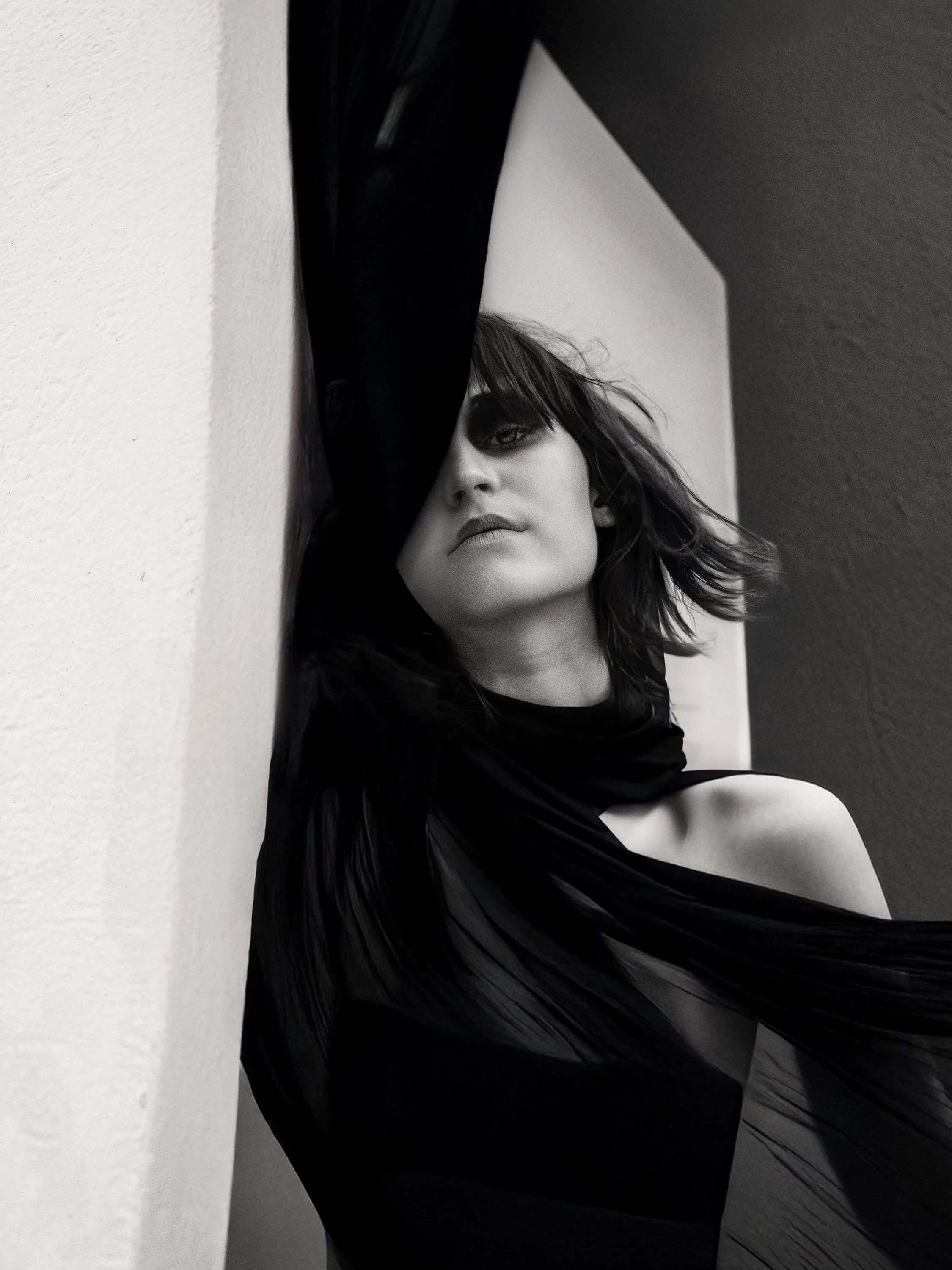 Black and white photo of a woman with dark hair, partially covering her face with one arm, looking at the camera, against a plain wall background.