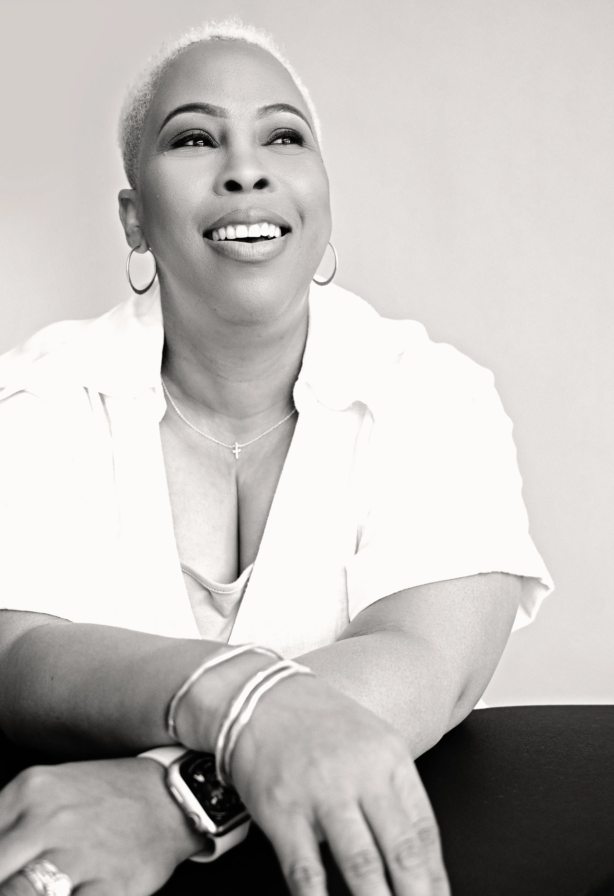 Black and white photo of a smiling woman with short hair, wearing hoop earrings, a necklace with a cross, and a white shirt. She has her arms crossed on a table or surface in front of her.