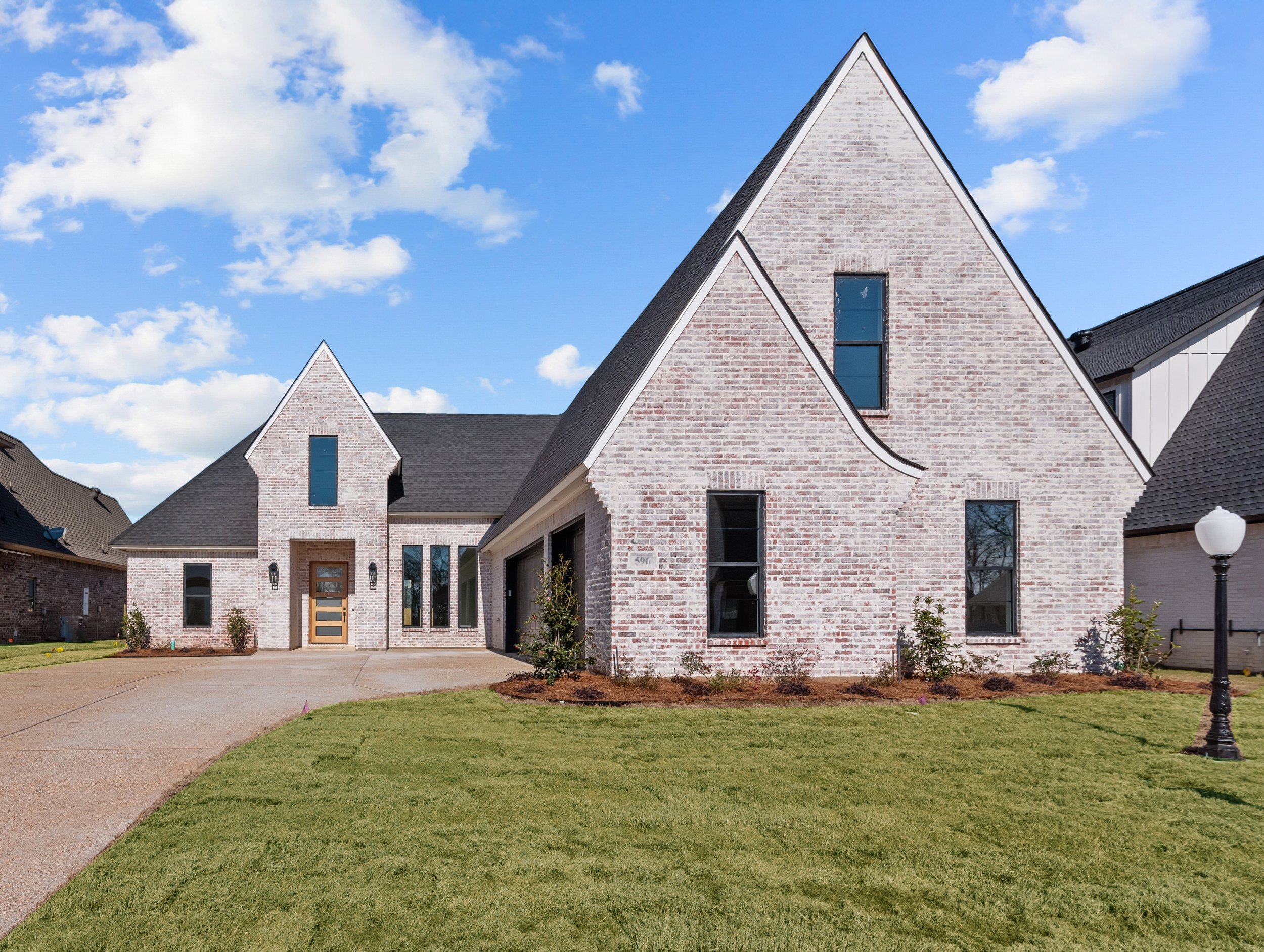 A brick house with a sharp triangular roof, a front porch with a wooden door, and a well-maintained lawn under a partly cloudy sky.