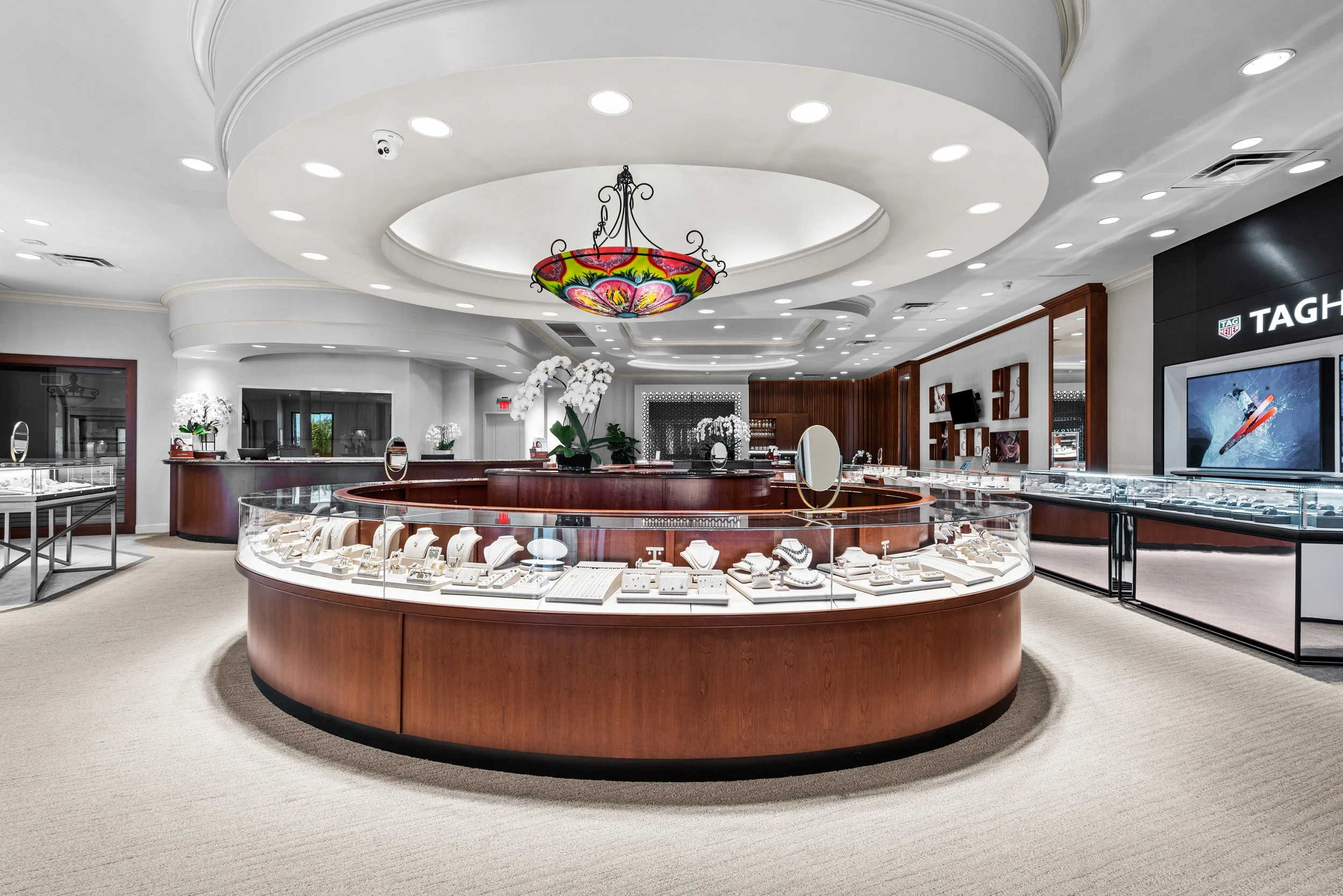 Jewelry display case inside a modern retail store or jewelry shop with a round glass counter filled with rings, necklaces, and other jewelry, surrounded by white orchids and a polished wood interior.