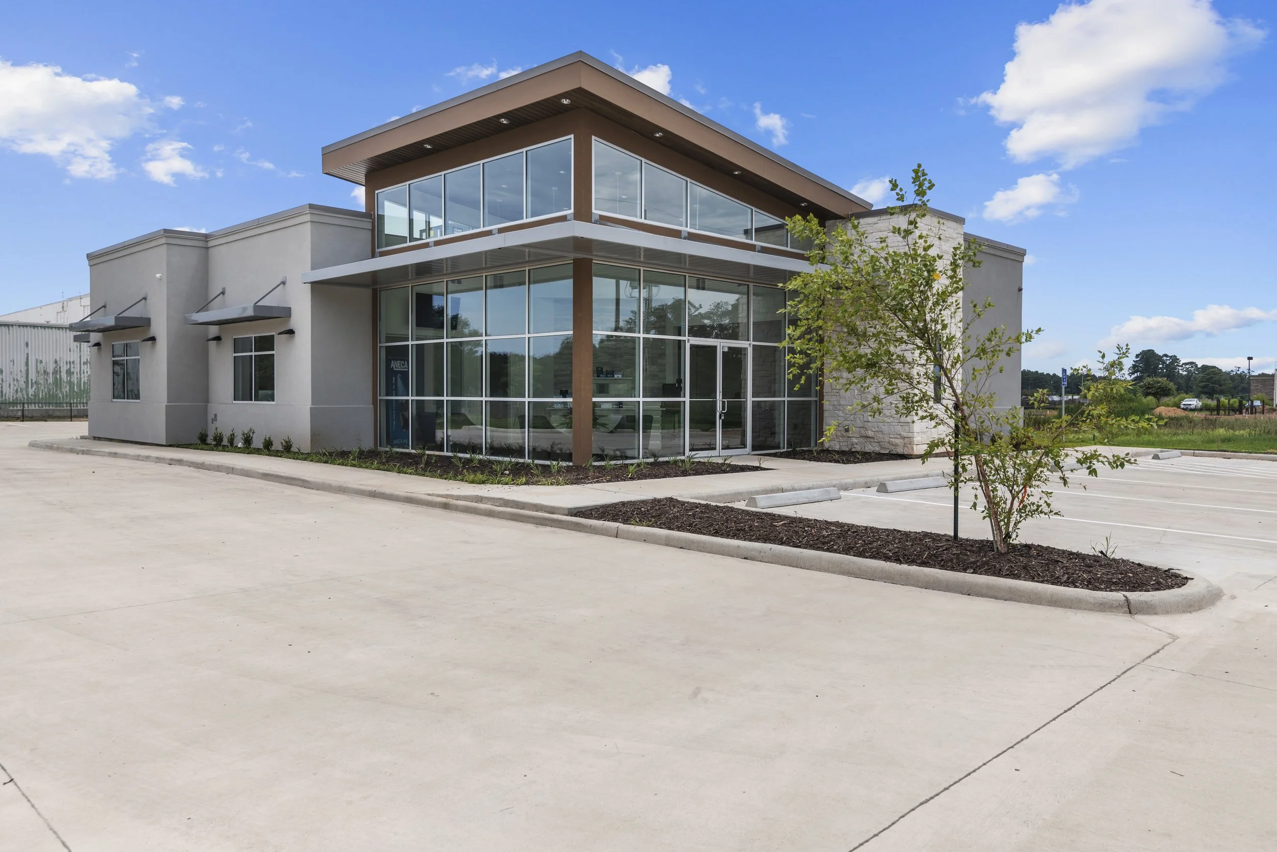 Modern commercial building with glass walls and a small tree in the foreground, parking lot in front, under a blue sky with some clouds.