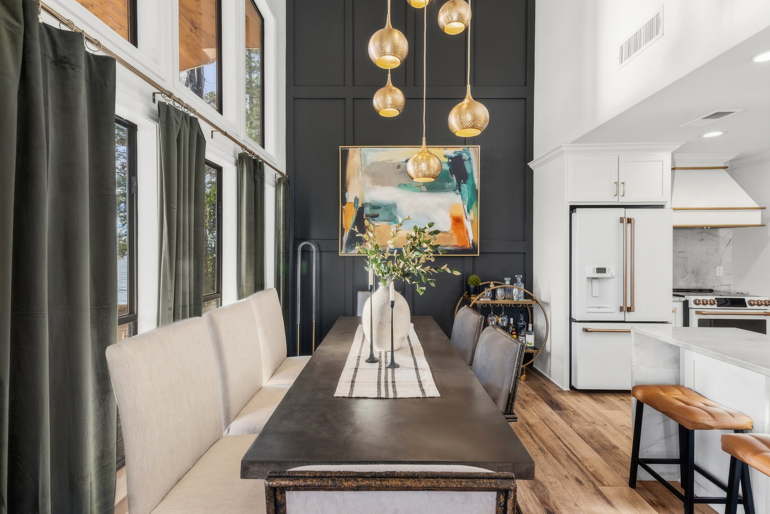 A modern dining room with a long dark wooden table, beige upholstered chairs, and a beige runner. There are two candle holders and greenery in a vase as centerpieces. Large windows with dark curtains on the left and a dark accent wall decorated with 