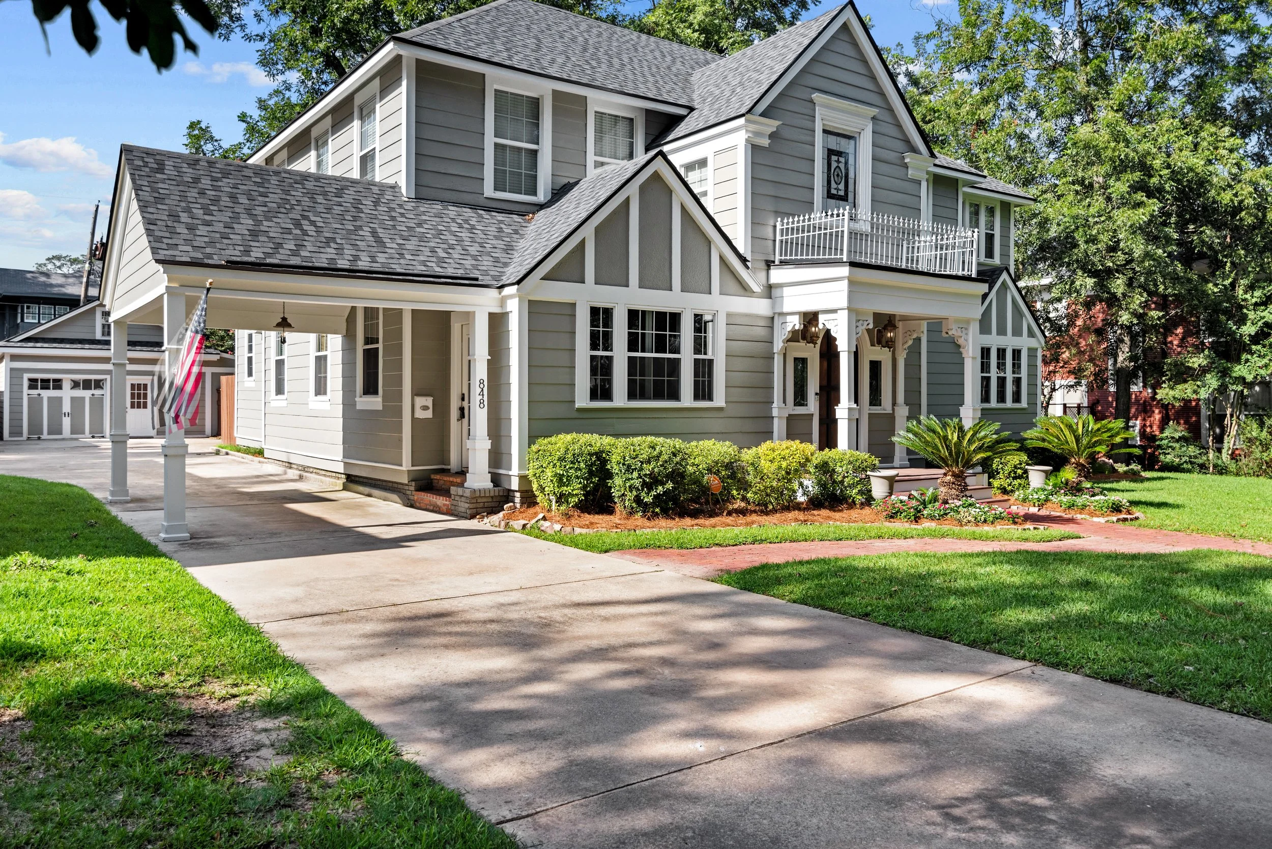 A two-story, gray and white house with a large front porch, surrounded by a green lawn and trees, with a concrete driveway leading to a garage, and a flagpole with an American flag in front.
