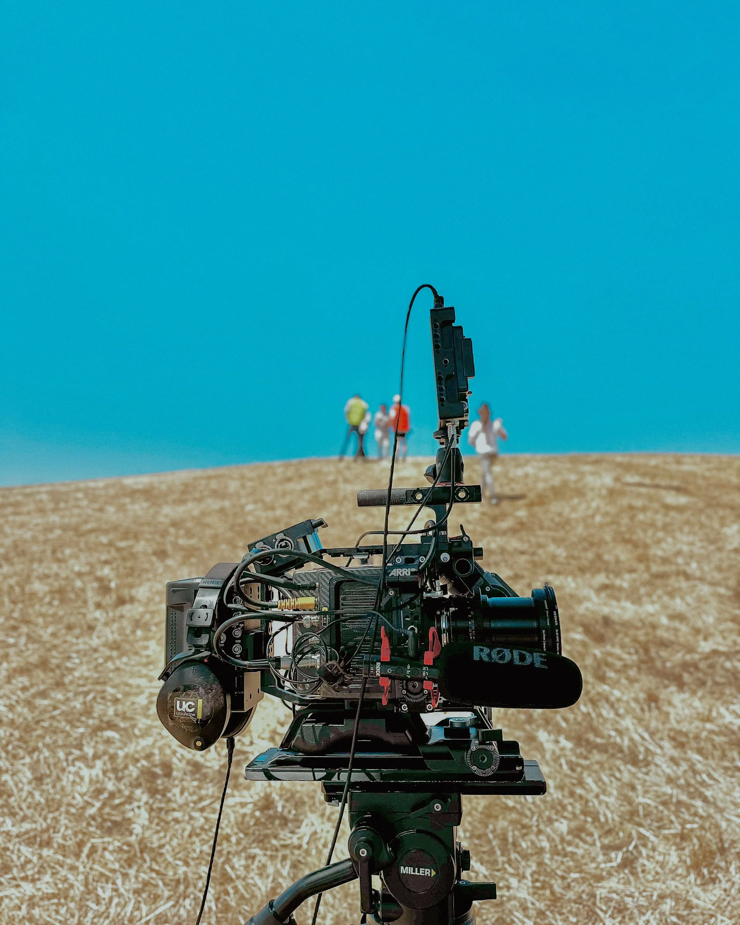 Close-up of a professional film camera set up outdoors on a tripod in a field, with a blurred group of people in the background under a clear blue sky.