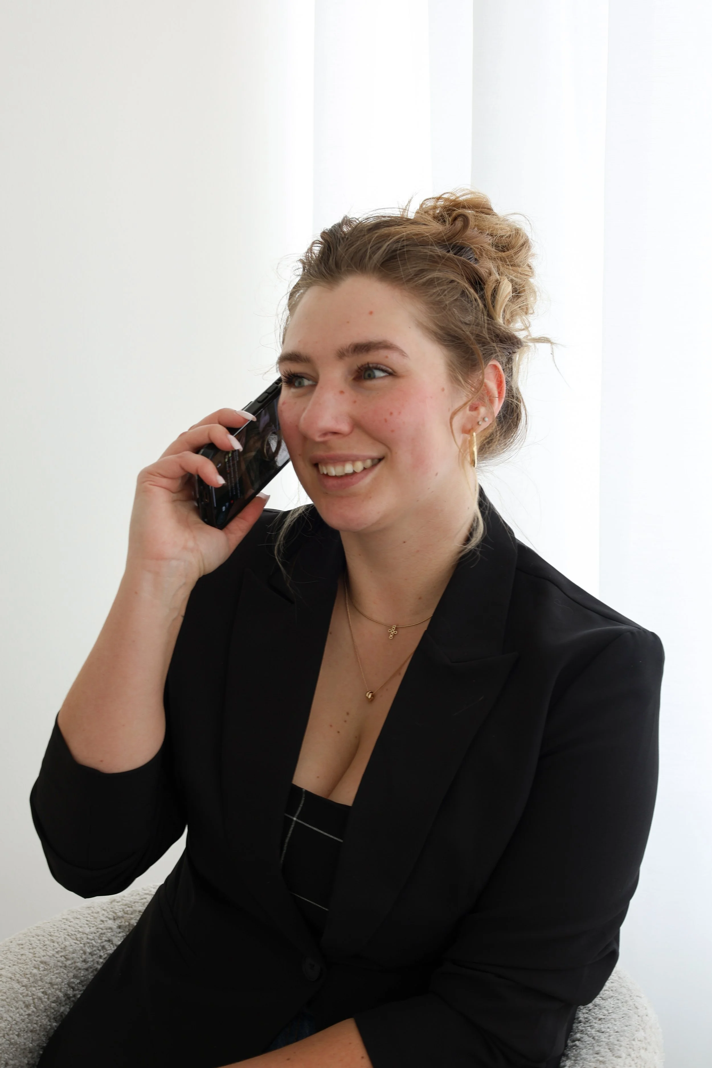 A young woman with curly hair in a bun, wearing a black blazer and jewelry, sitting on a chair, smiling and talking on her phone.