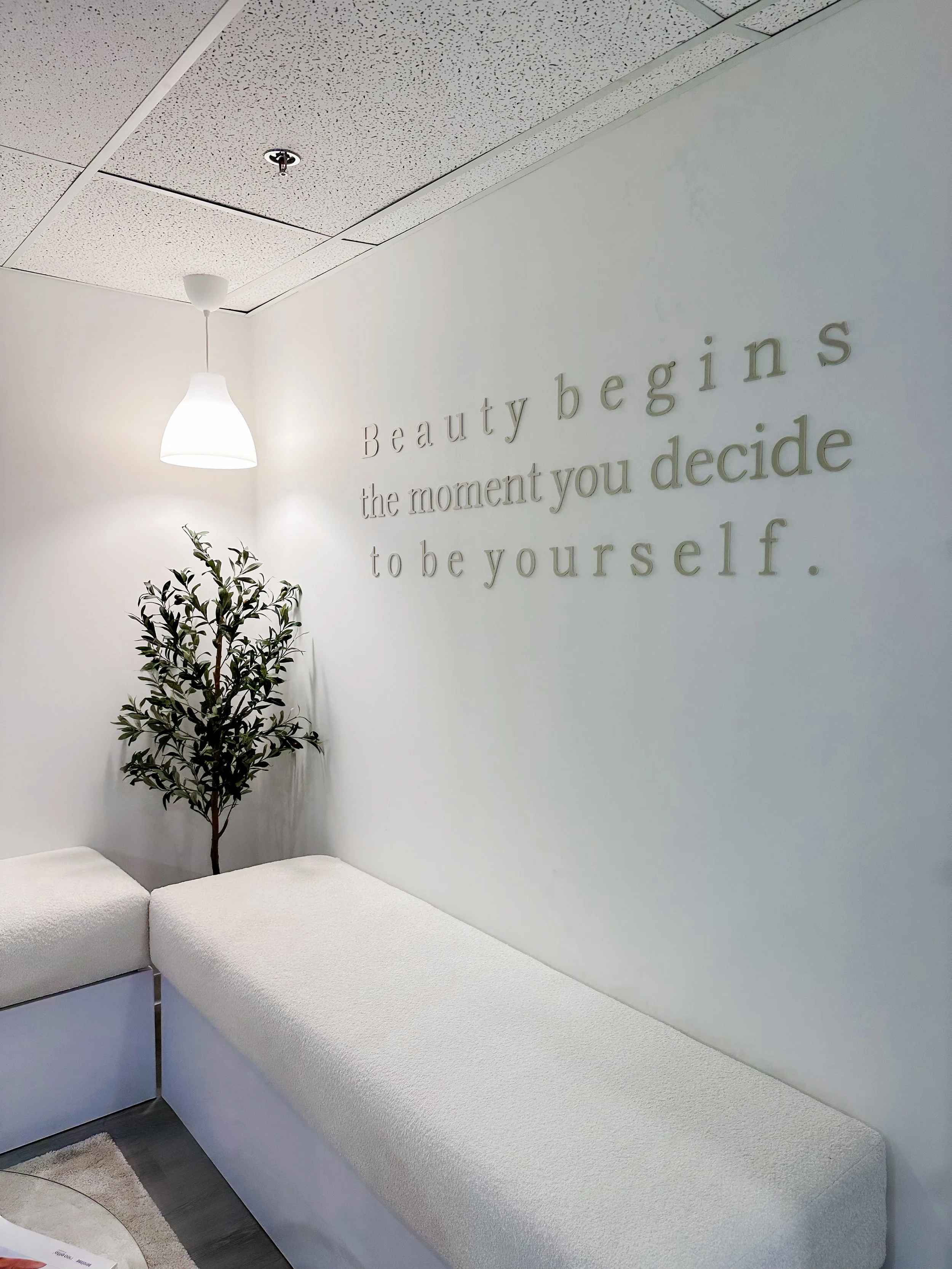 A white wall with the quote 'Beauty begins the moment you decide to be yourself' in raised letters, a small potted tree, a white hanging lamp, and beige seating.