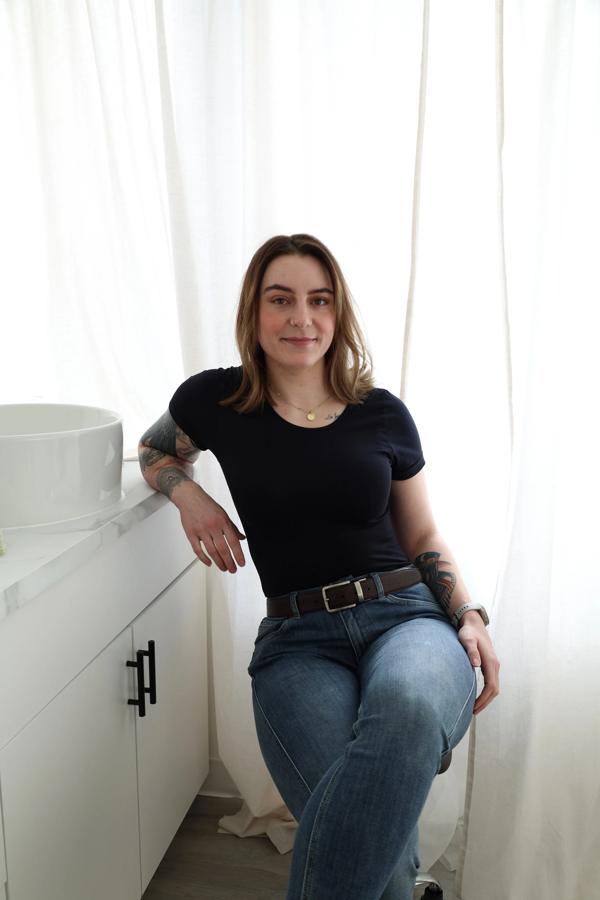 A woman with shoulder-length brown hair, wearing a black t-shirt and blue jeans, sits on a stool next to a white countertop with a large white bowl on it, in front of white curtains in a well-lit room.