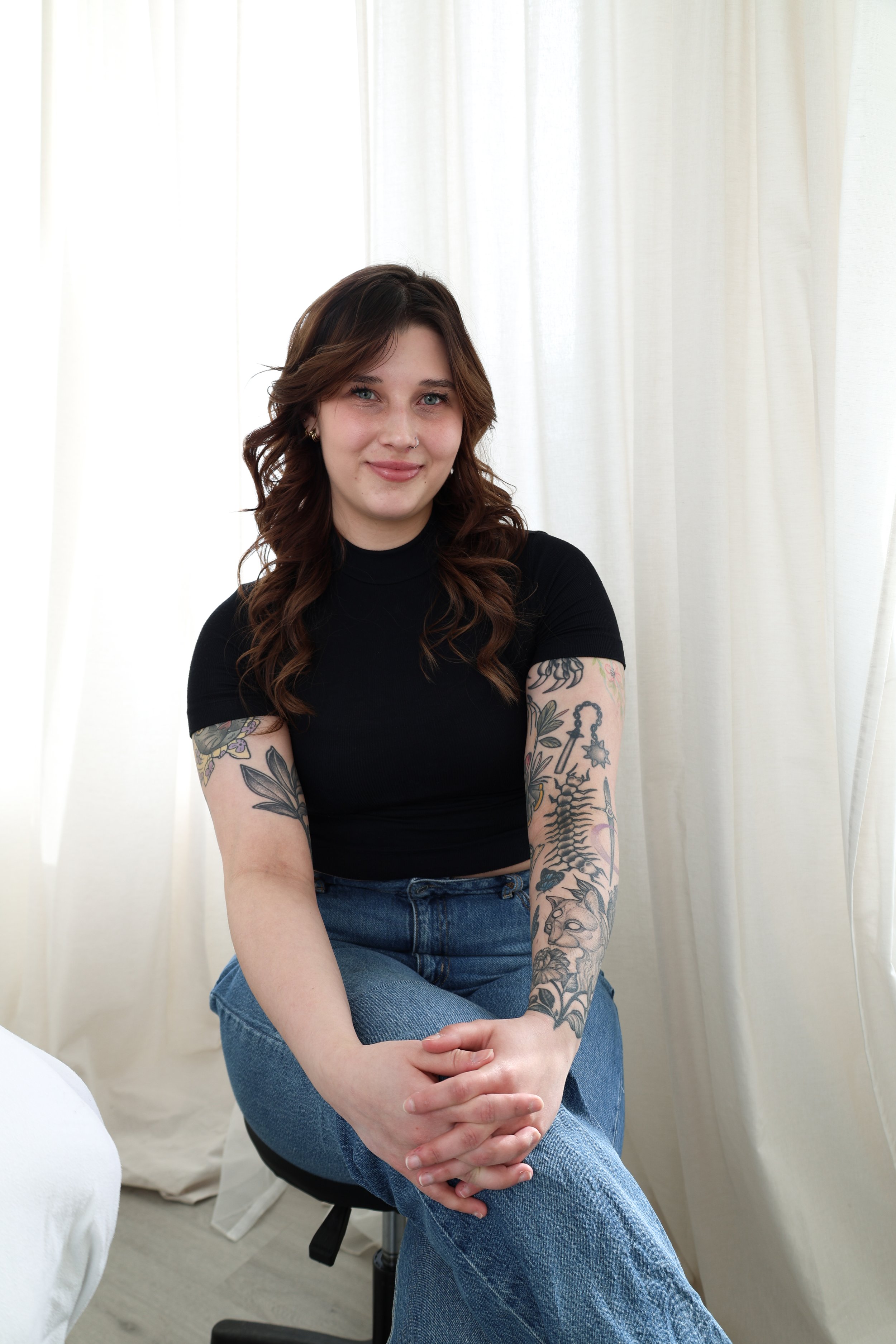 A young woman with curly brown hair, tattoos on both arms, wearing a black shirt and blue jeans, sitting on a black stool with her hands clasped, smiling softly at the camera, in front of white curtains.