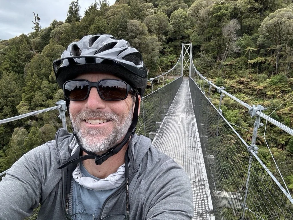 Matt Freeman taking a selfie on a suspension bridge in a lush green forest in New Zealand, wearing a bicycle helmet and sunglasses, smiling at the camera.
