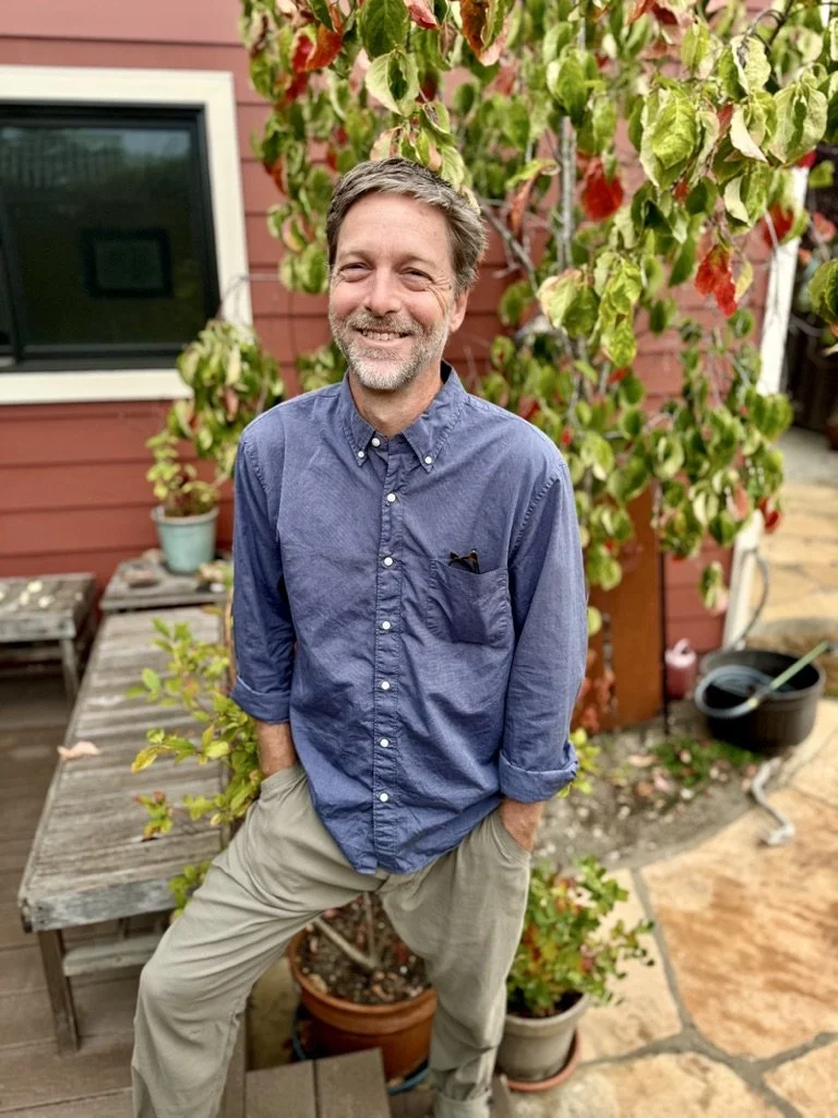 Matt Freeman stands outdoors in front of a Cherokke dogwood, with a wooden deck, potted plants, a red house wall, a window, and garden tools nearby.