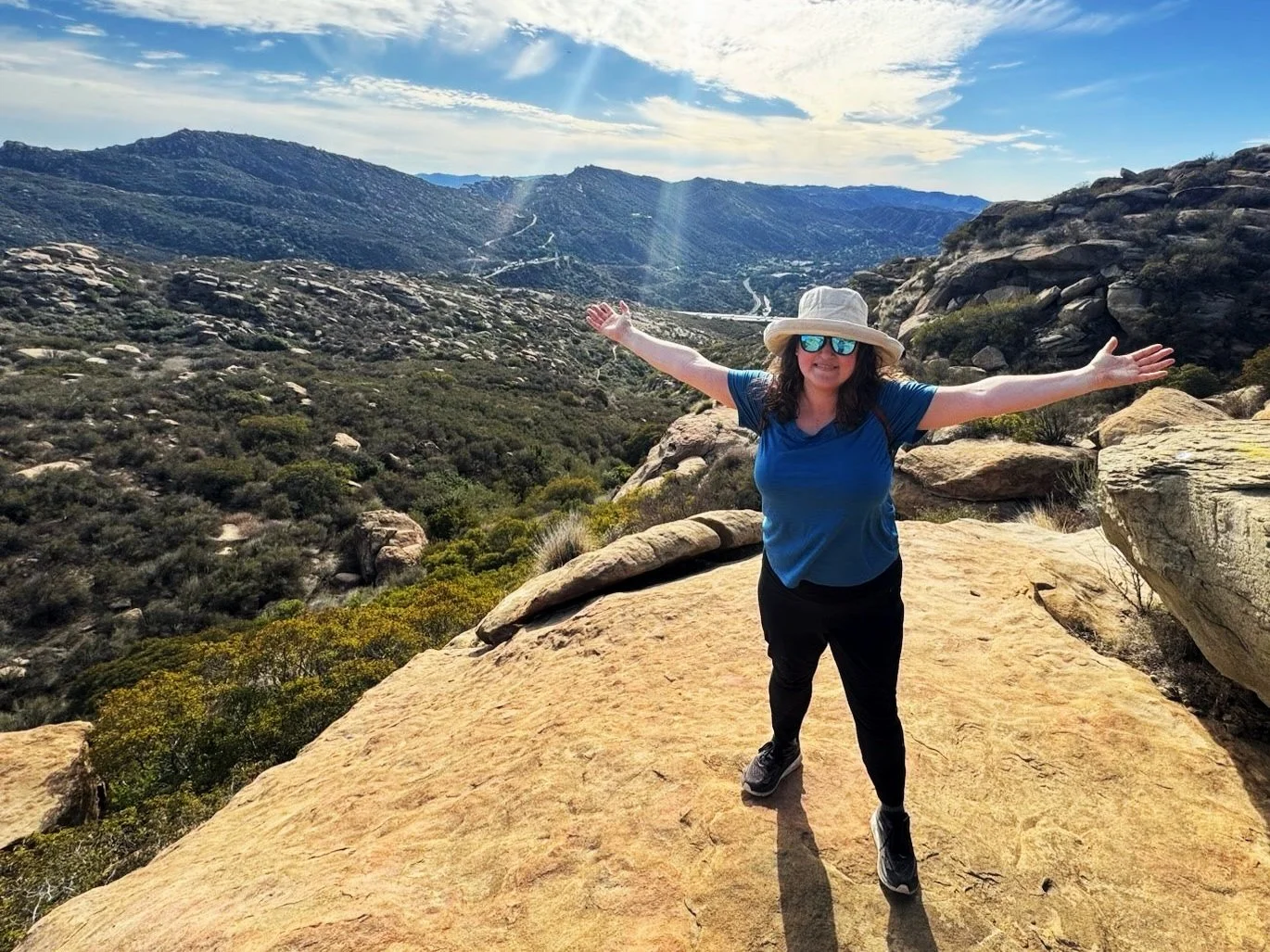 A woman in a blue shirt, black pants, and a wide-brimmed hat stands on a large rock with her arms outstretched in a mountainous desert landscape, under a partly cloudy sky.