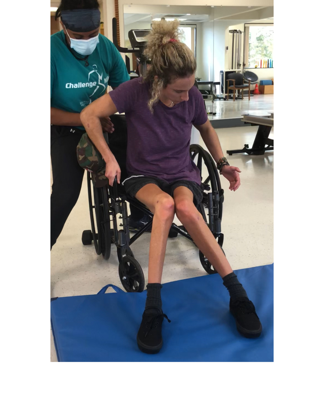 A woman in a wheelchair performing a physical therapy exercise while a healthcare professional supervises. The woman is on a blue exercise mat, with her legs extended and feet in black shoes, wearing a purple shirt and black shorts. The healthcare professional, wearing a teal shirt, black pants, and a face mask, supports her from behind in a well-lit physical therapy room.