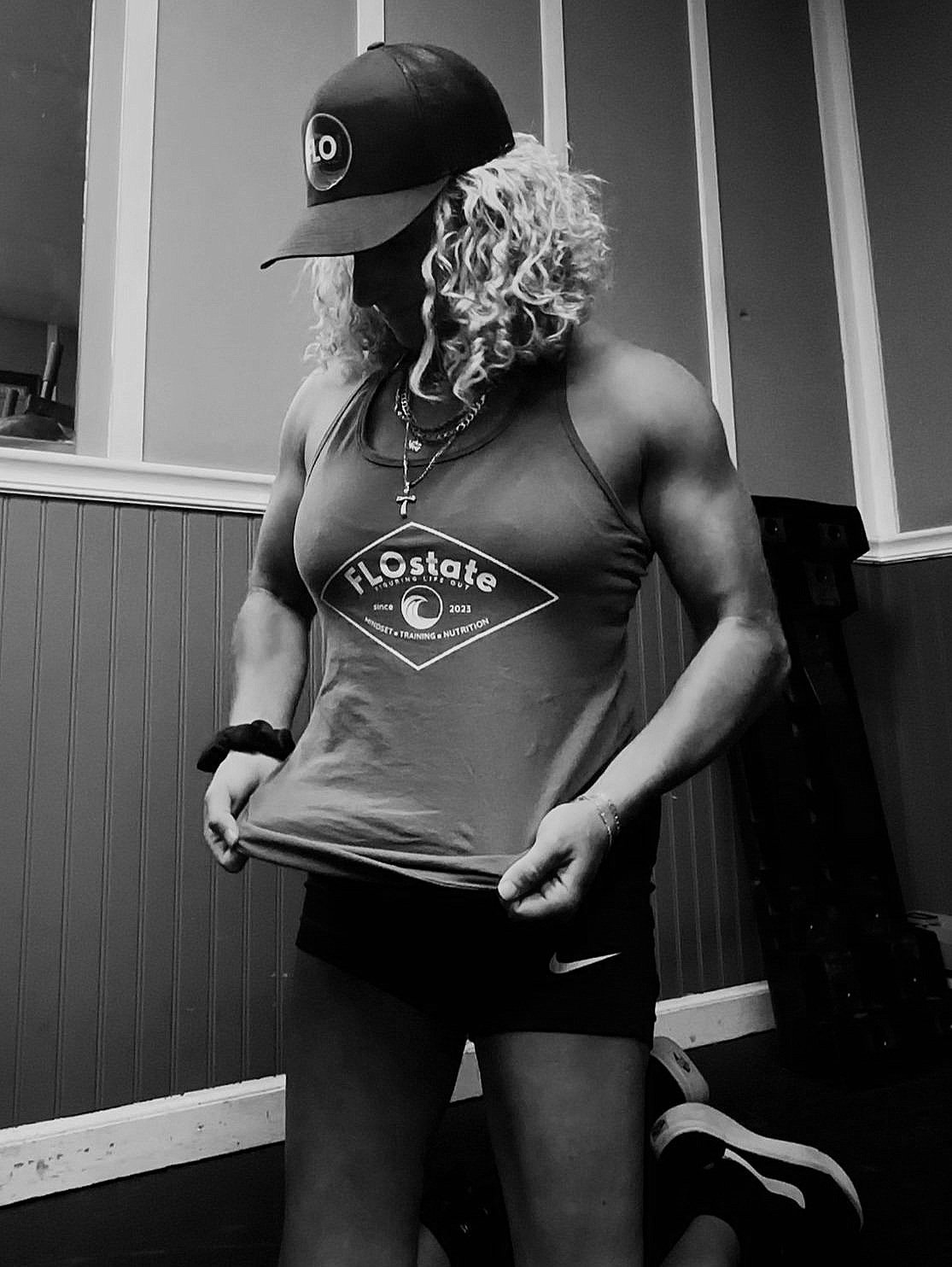 A woman with curly blonde hair roller-skates indoors, wearing a tank top with 'FLO State' logo, shorts, a cap, necklaces, and a bracelet, with her head down and hands pulling at her shirt.