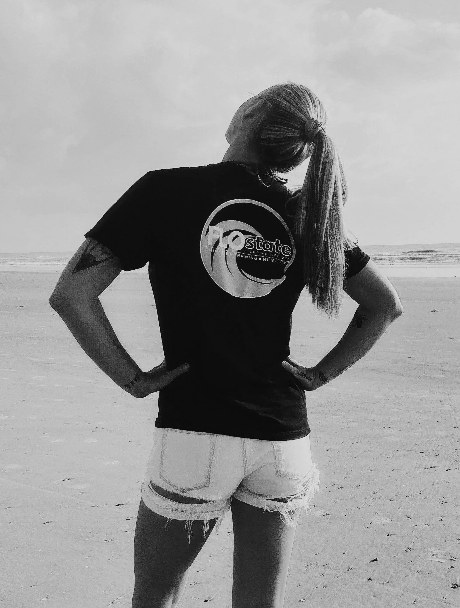 A woman with long hair in a ponytail standing on a beach with her back to the camera, hands on hips, wearing a T-shirt and ripped denim shorts, looking out at the ocean.