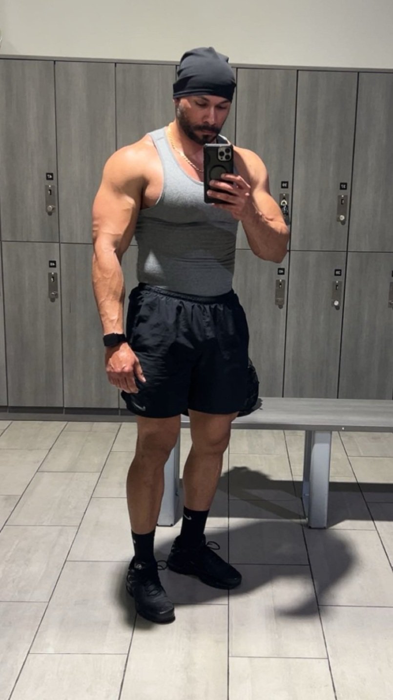 A muscular man in a gray tank top, black athletic shorts, black socks, and shoes takes a mirror selfie in a locker room, wearing a dark beanie.
