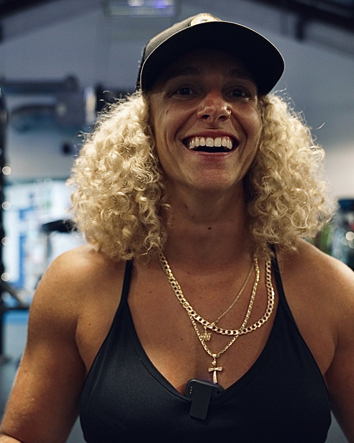 Smiling woman with curly blonde hair wearing a black tank top, gold necklaces, a black hat, and a nose ring, in a gym setting.