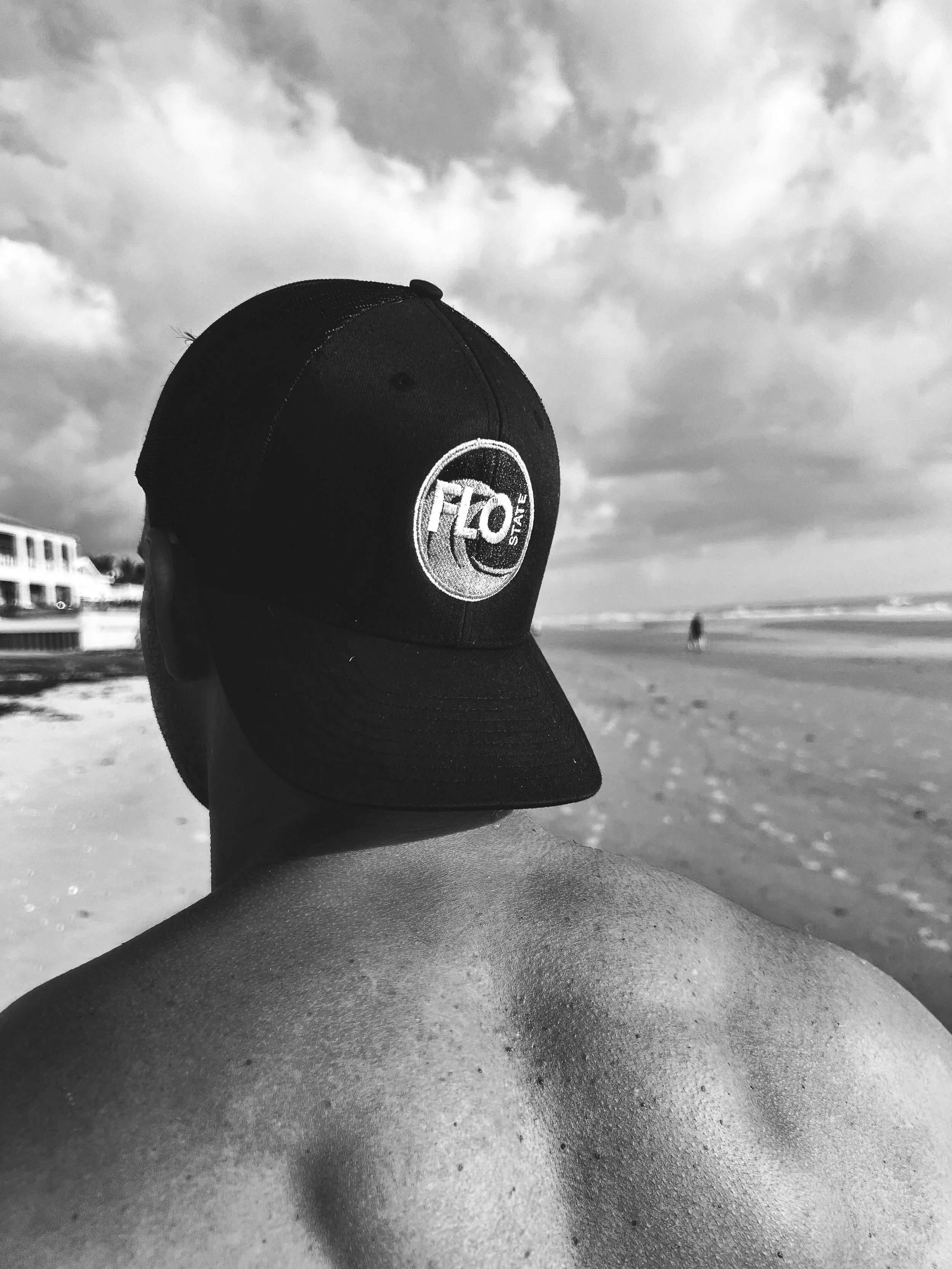 Back view of a person on the beach wearing a black cap with a logo, with sand and ocean in the background.