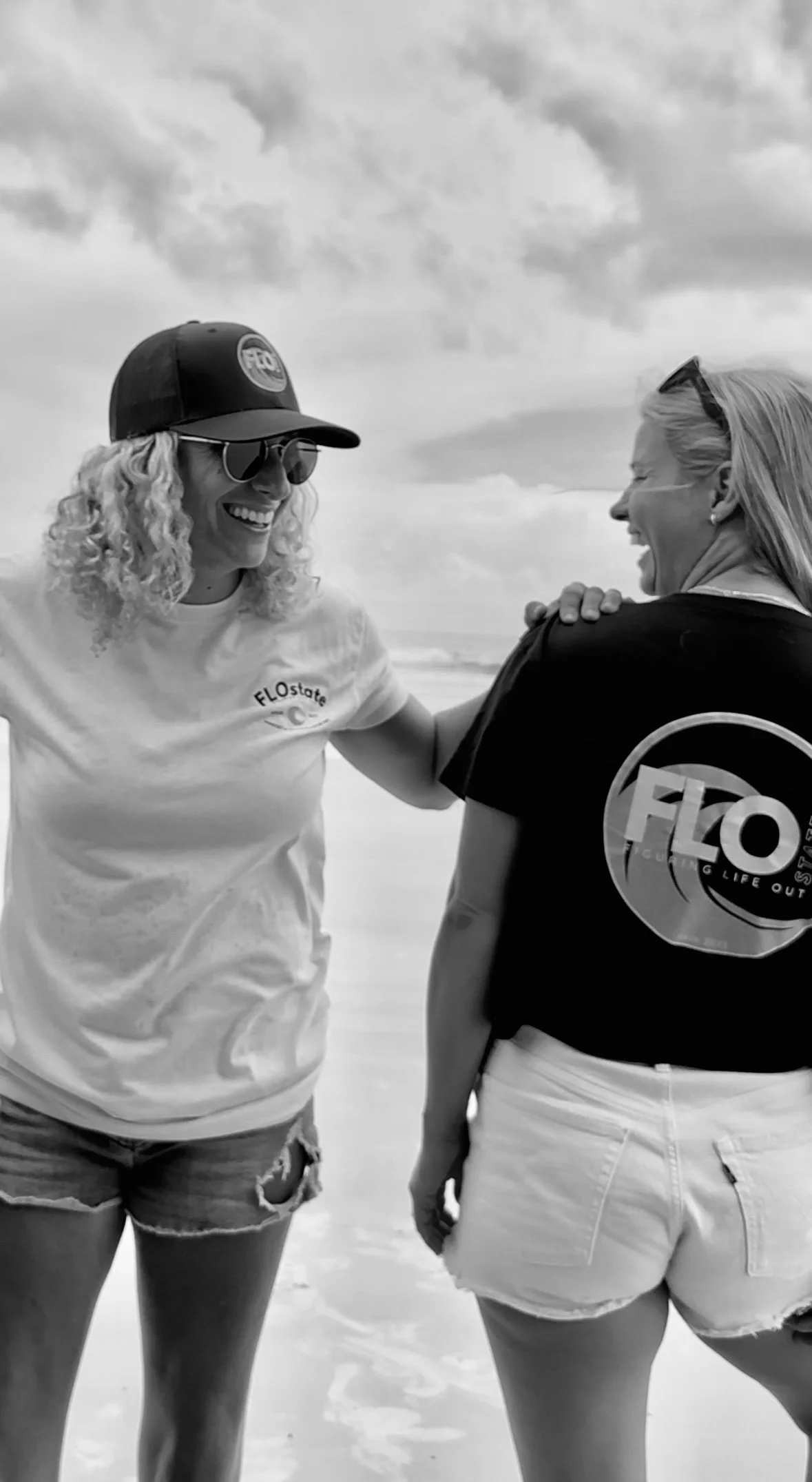Two women at the beach, smiling and laughing, engaging in conversation. One woman with curly hair and sunglasses wearing a cap and T-shirt, the other with straight hair and sunglasses on her head, wearing a T-shirt and shorts. The cloudy sky is visible in the background.