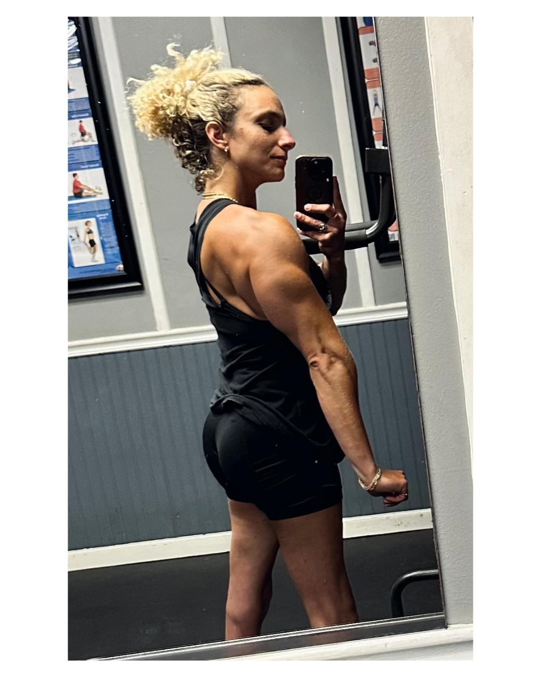 Woman with curly hair taking a mirror selfie in a gym, wearing black workout clothes.
