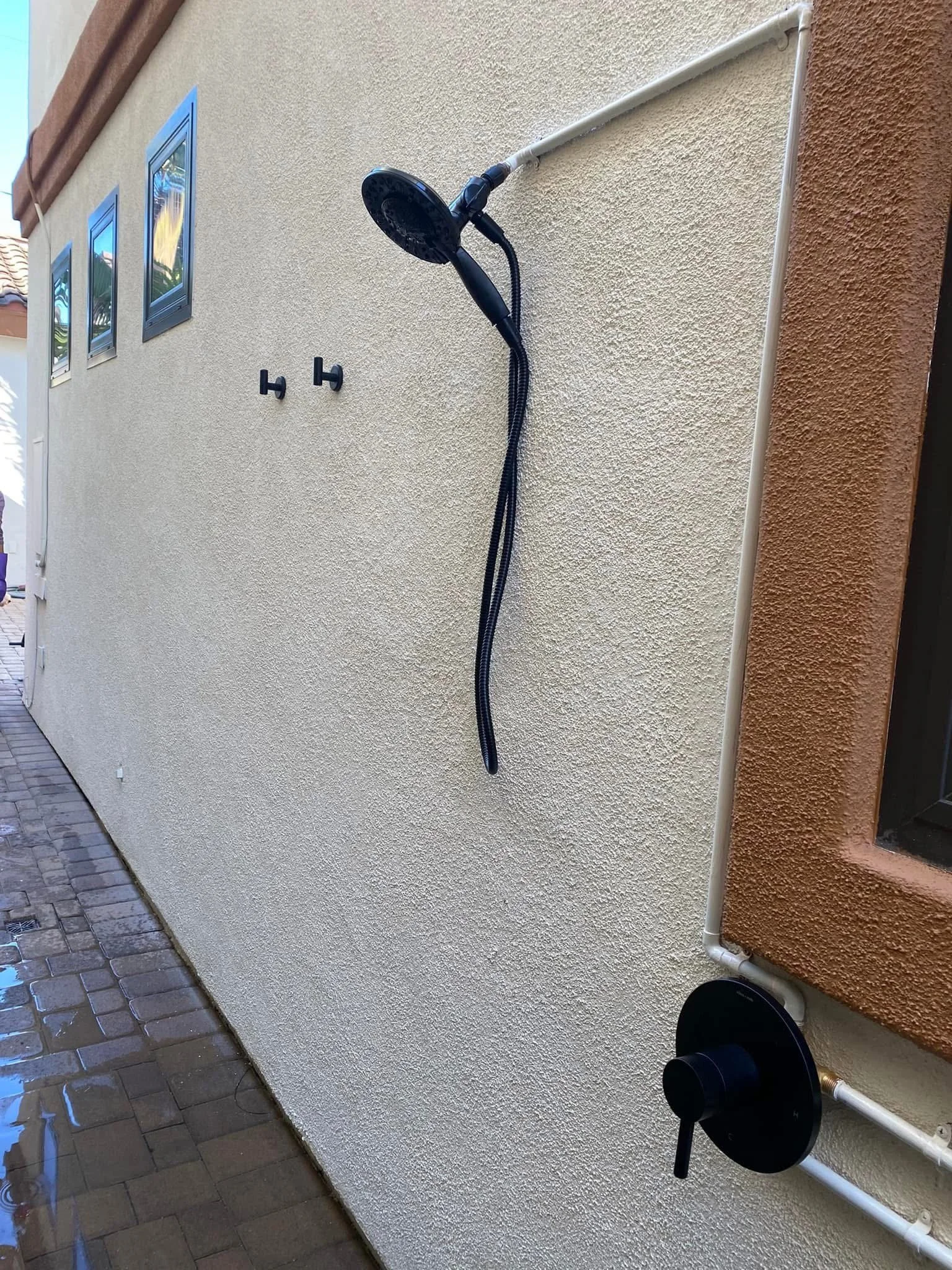 An outdoor shower with a black showerhead and hose mounted on a beige stucco wall, with a white pipe and two small windows above and a mudroom door to the side.