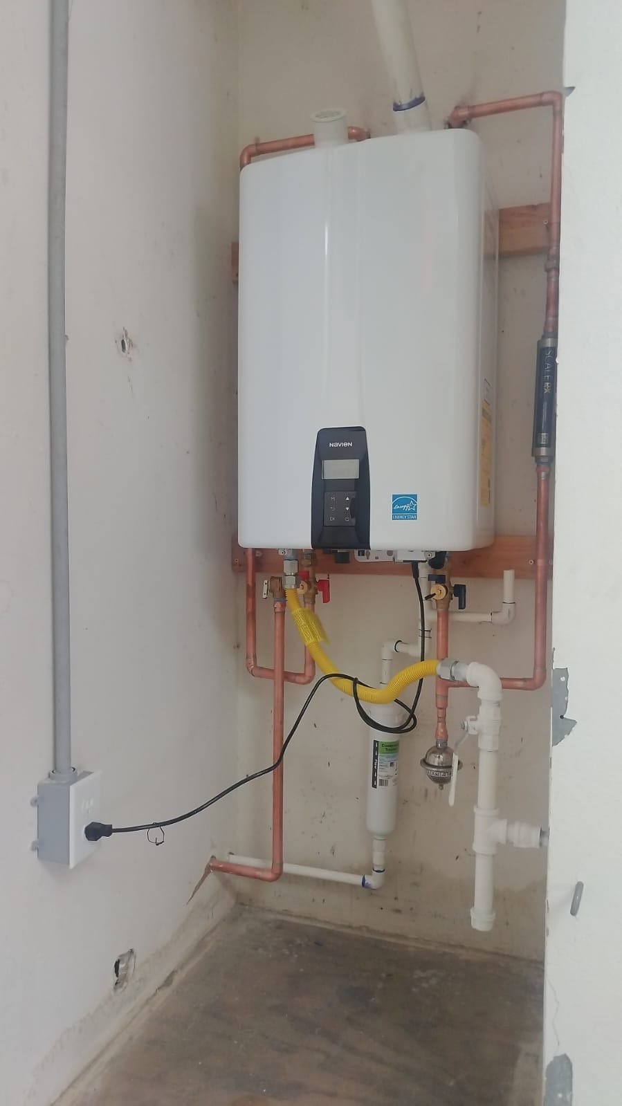 A tankless water heater installed on a wall with copper and PVC pipes, and electrical wiring, in an unfinished or utility room.