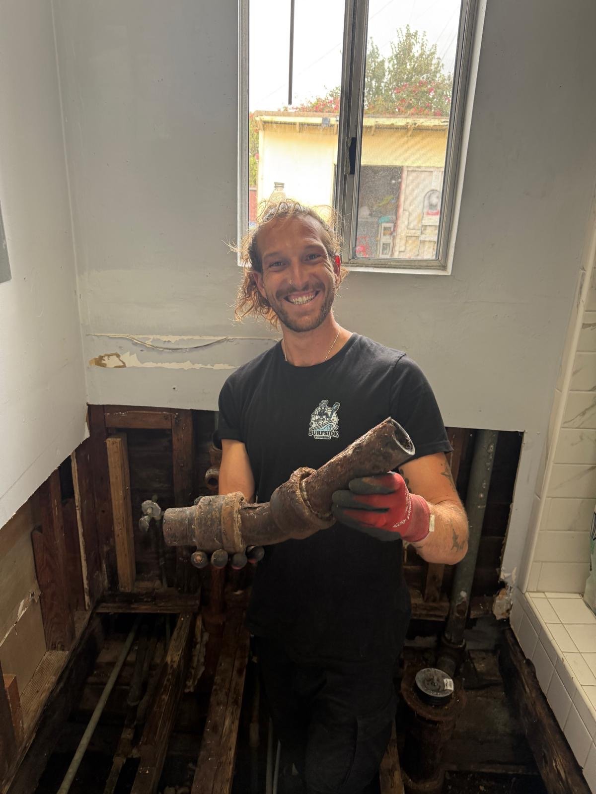 A man smiling, wearing gloves, holding a rusty pipe inside a partially renovated room with exposed wooden framing.