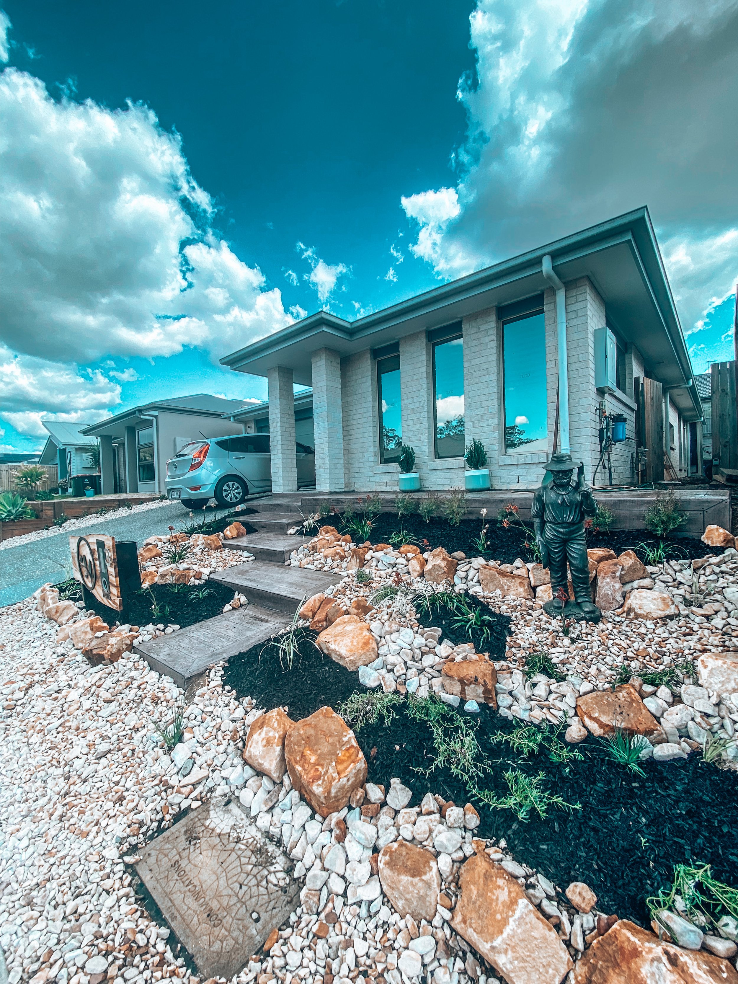 Front yard garden with rocks, plants, and a small statue of a man, in front of a modern house with large windows and a car parked in the driveway.