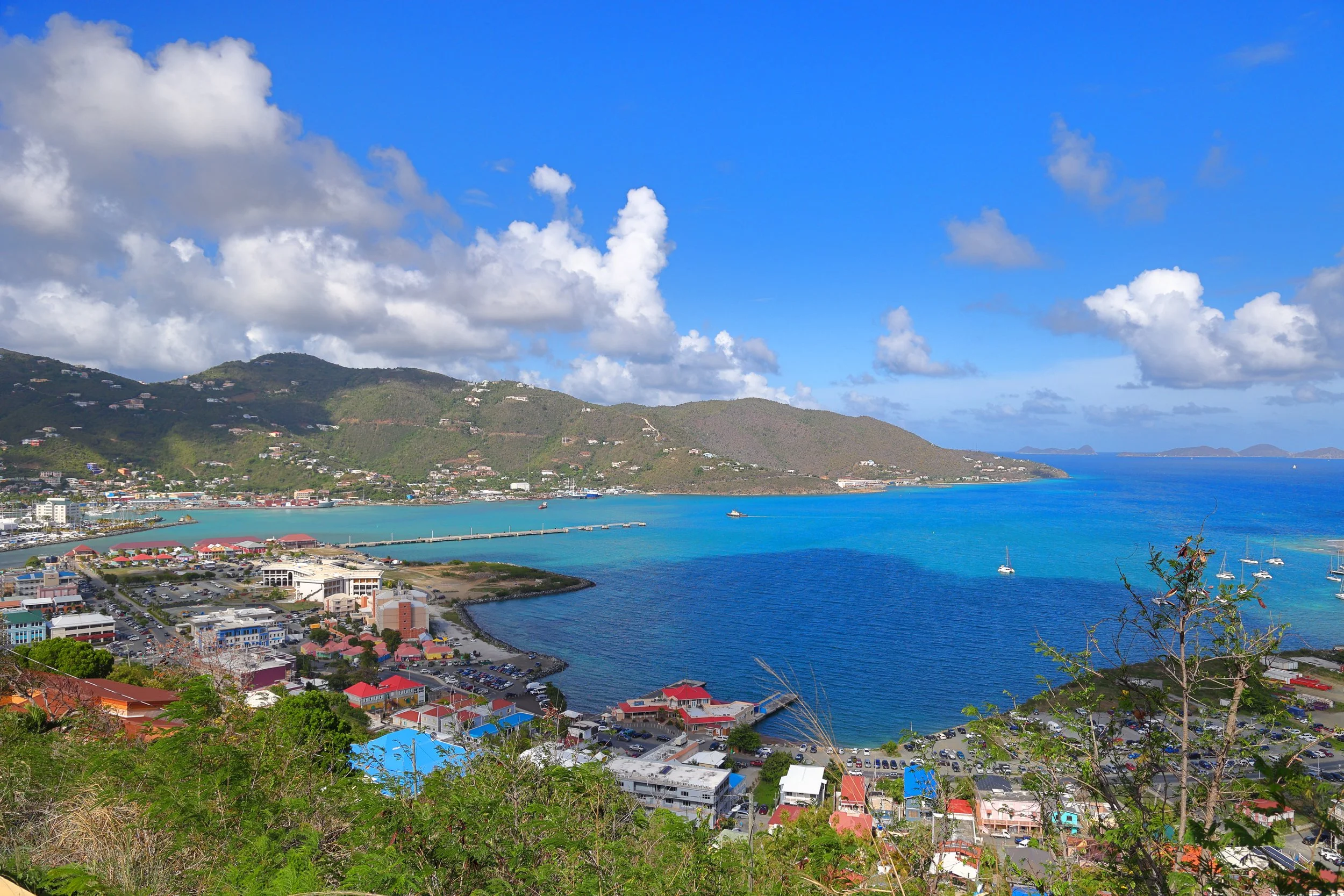 A panoramic view of a Caribbean coastal town , road town, with vibrant buildings, marina with sailboats, and lush green hills under a partly cloudy blue sky.