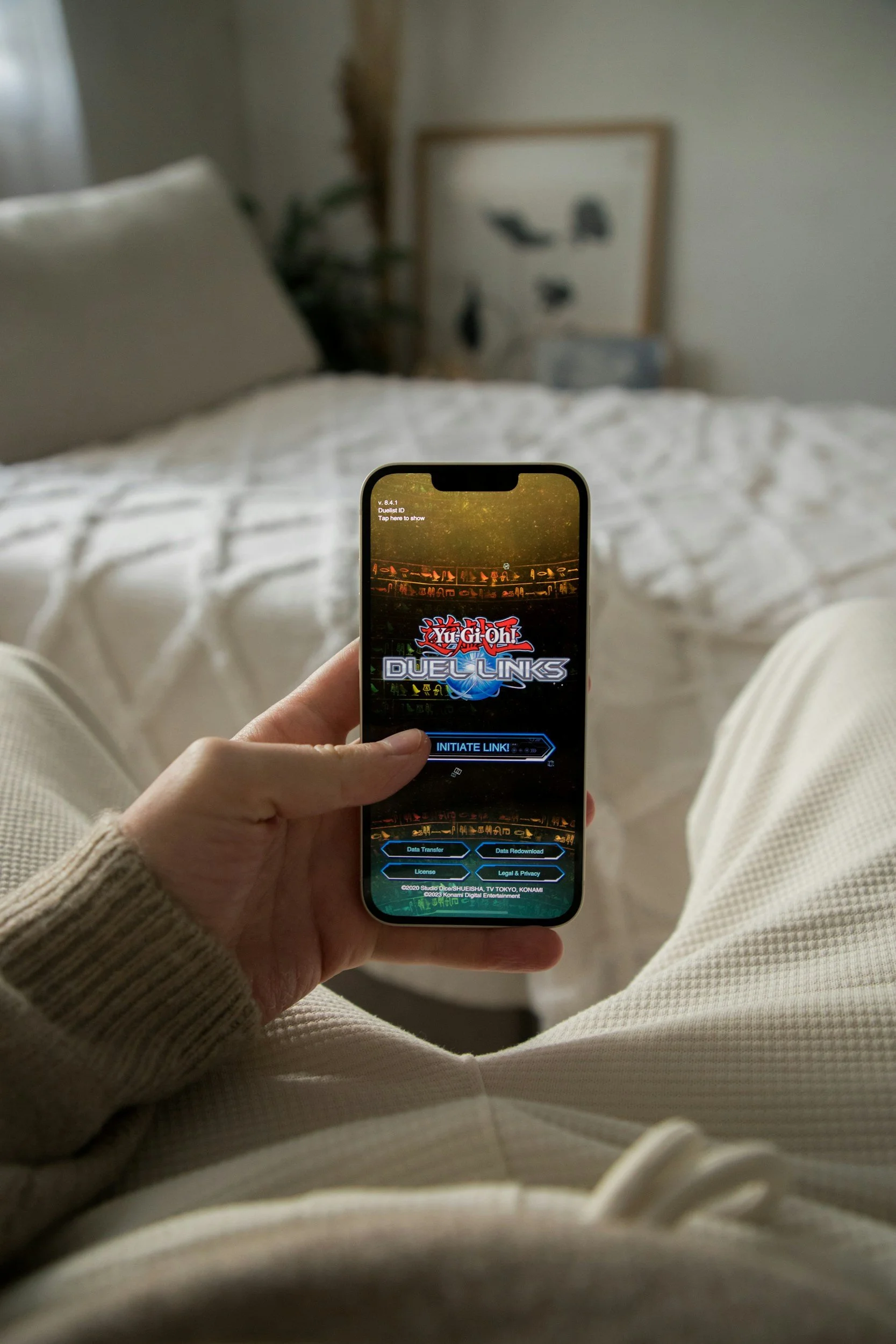 Person sitting on a bed playing the mobile game Yu-Gi-Oh! Duel Links on their smartphone.