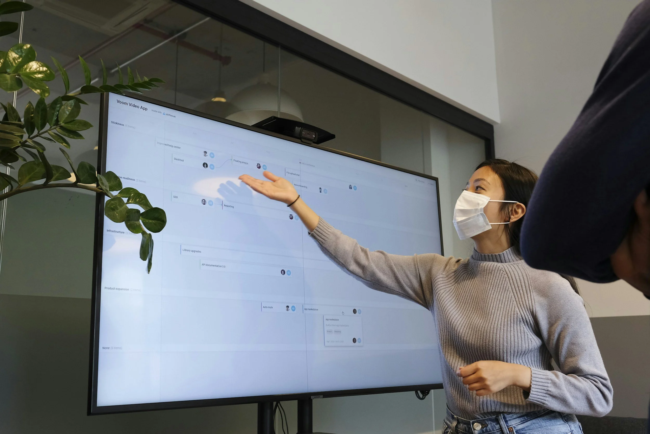 A woman wearing a face mask and beige sweater is pointing at a large screen displaying a project management or planning app, with another person partially visible in the foreground.