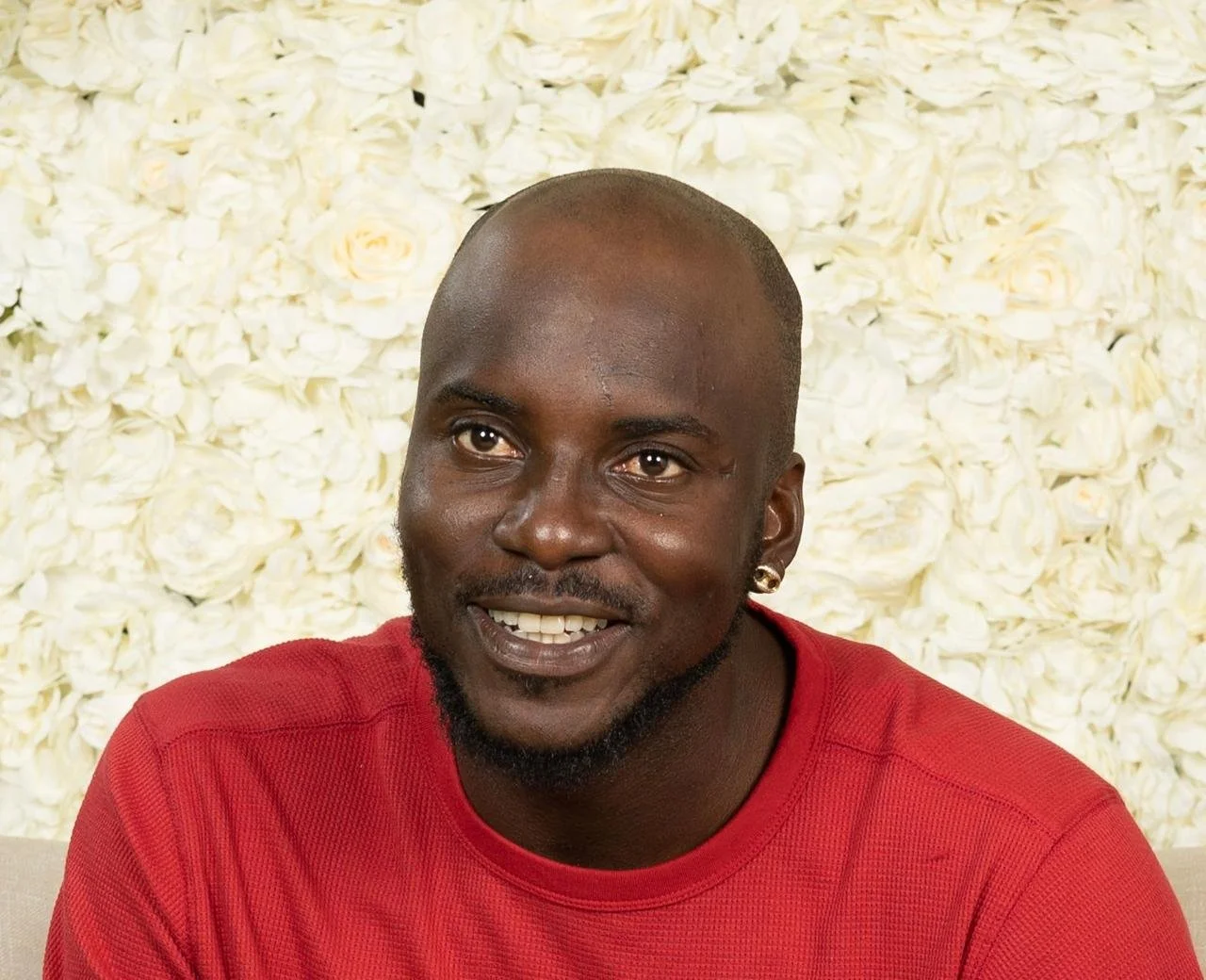 A smiling Black man with a shaved head and a beard, wearing a red shirt and a gold earring, in front of a cream-colored floral background.