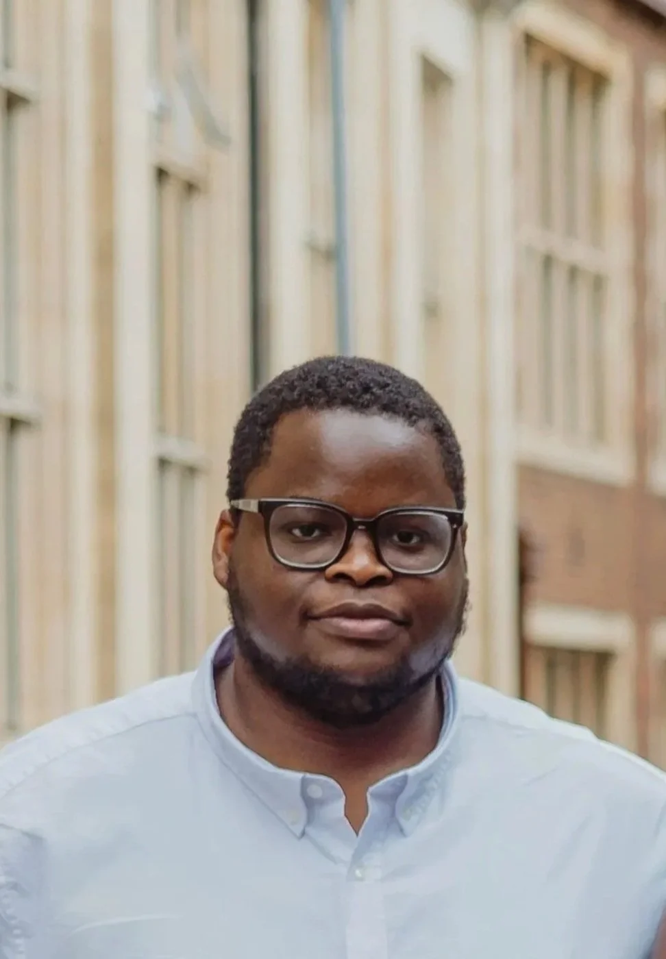 A young man with glasses and facial hair wearing a light blue shirt standing in front of a background of bank vault doors.