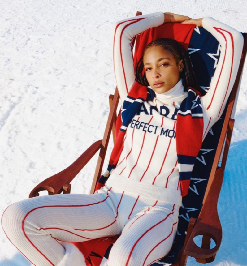 A young woman relaxing on a wooden lounge chair outdoors on snow, wearing a coordinated red, white, and blue striped sweater and pants, with a matching scarf and a white turtleneck underneath.