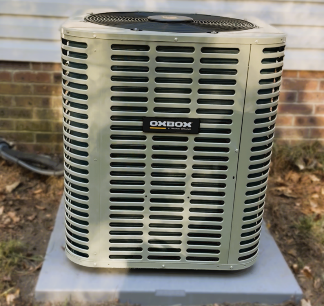 Outside view of a beige outdoor air conditioning unit with black vents, placed on a gray concrete pad in front of a house with white siding and a brick foundation.