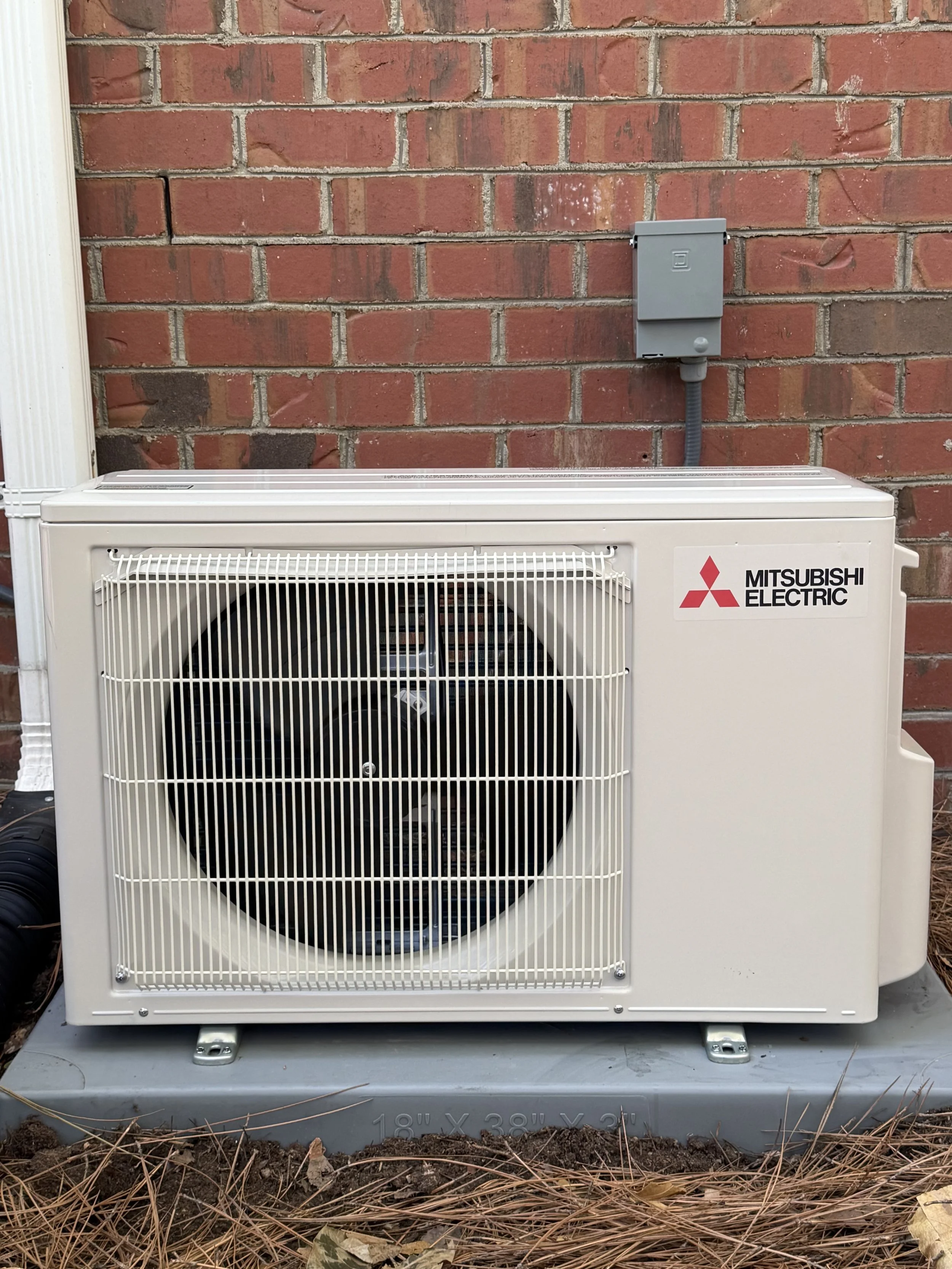 An outdoor Mitsubishi Electric heat pump or air conditioning unit installed against a red brick wall, with a gray electrical box above it and some dried pine needles in front.