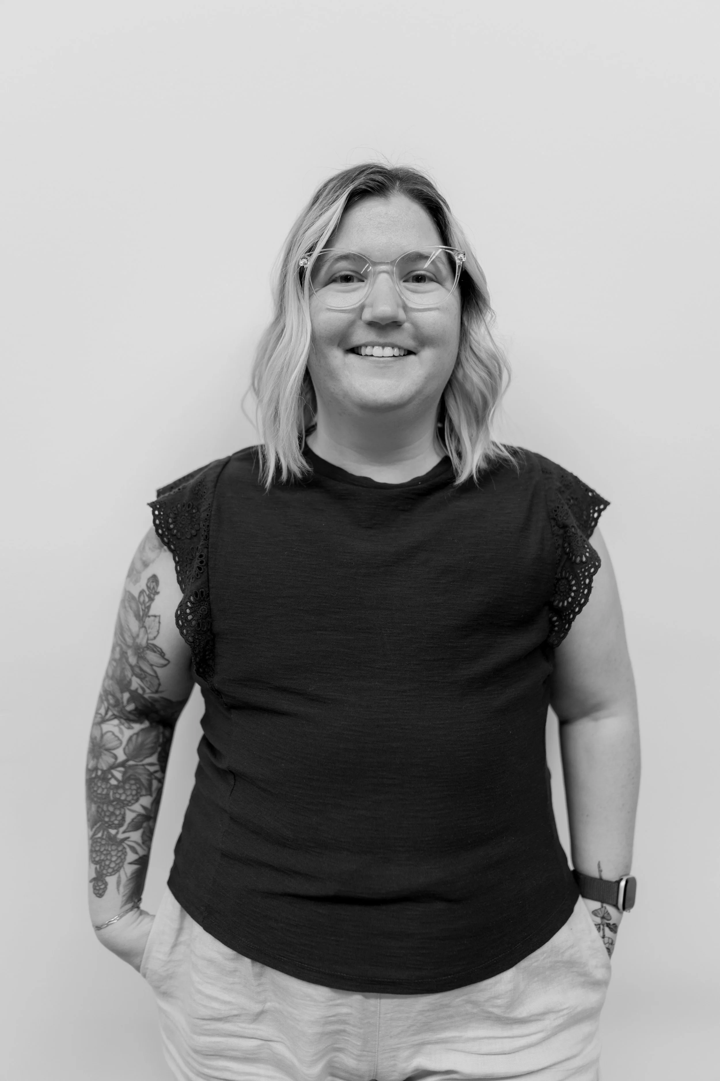 Black and white portrait of a smiling woman with glasses, wavy shoulder-length hair, tattoos on her left arm, wearing a black top with lace sleeves, standing against a plain background.