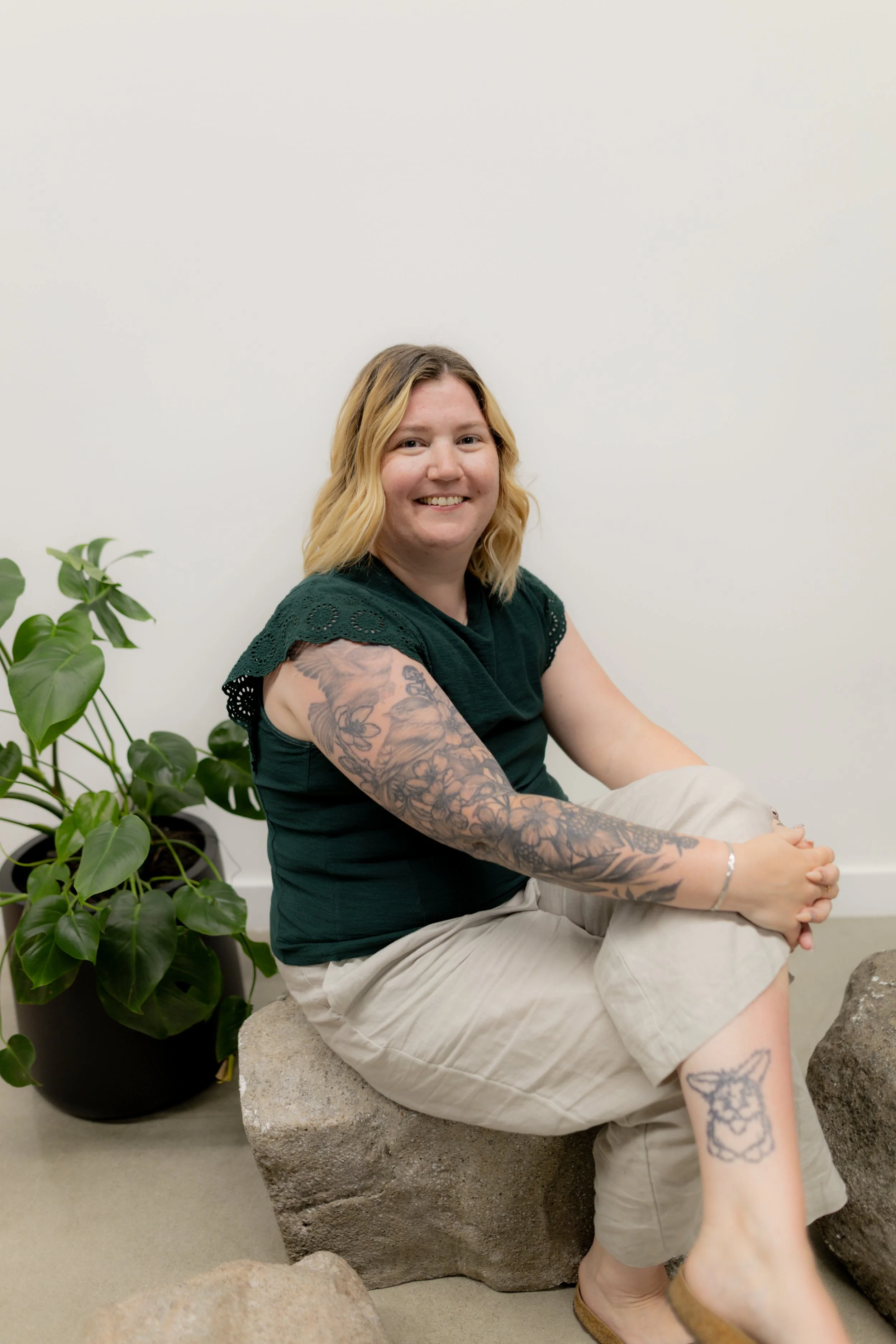 A woman with blonde hair and tattoos sitting on a rock, smiling at the camera, beside a green potted plant against a plain white wall.