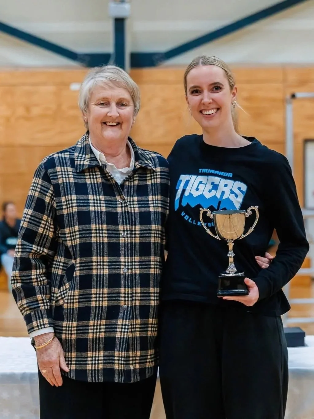 A young woman in a black sports jersey holding a trophy, standing next to an older woman in a plaid shirt, both smiling inside a gymnasium.
