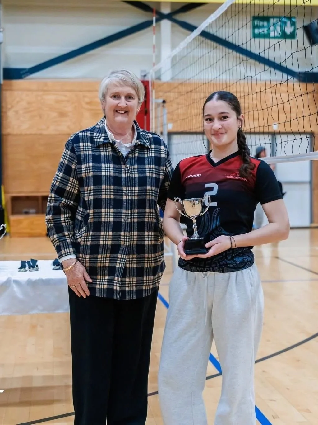 A young woman holding a trophy stands beside an older woman in a gymnasium with a volleyball net. The young woman is wearing a volleyball jersey and sweatpants, smiling, and the older woman is wearing a plaid jacket and black pants, also smiling.