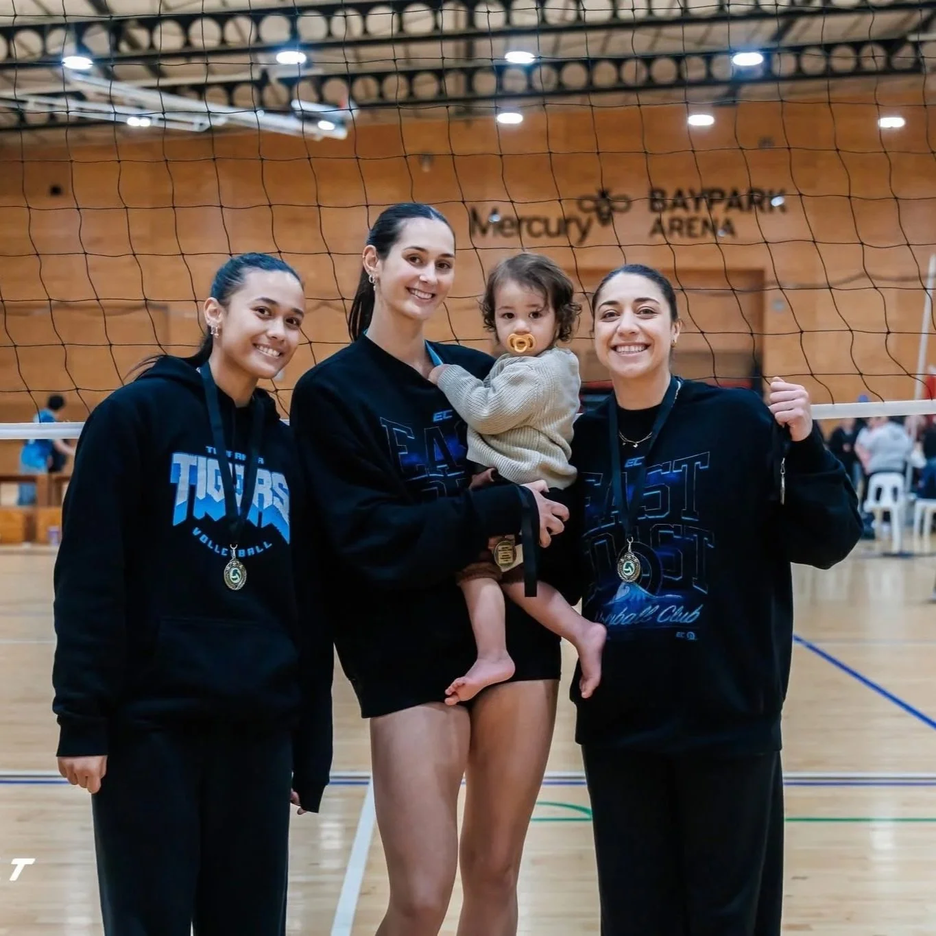 Four women and a young child at a volleyball court. Three women are wearing black sweatshirts, and one woman is holding a child with a pacifier. They are smiling and have medals around their necks. The background shows a wooden wall with signage for Mercury Baypark Arena.