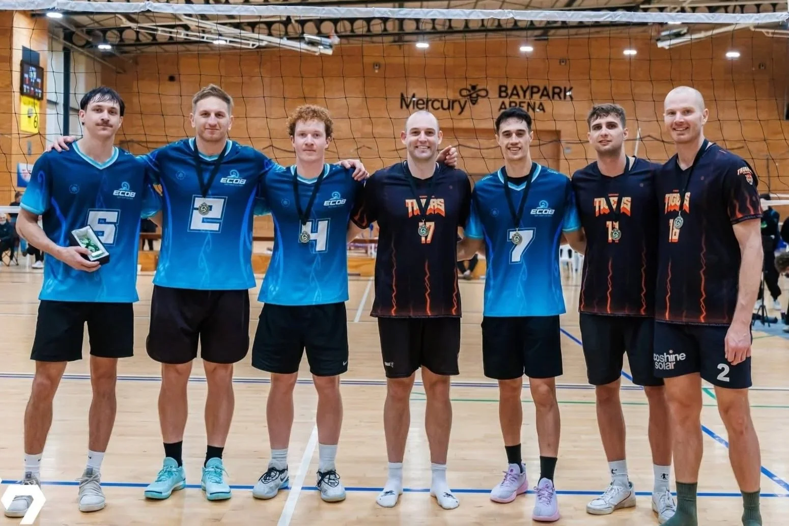 A group of seven male volleyball players standing in a line on an indoor volleyball court, wearing medals around their necks and their team uniforms. They are smiling and posing for a team photo after a match or tournament, with a volleyball net and the Mercury Baypark Arena signage in the background.