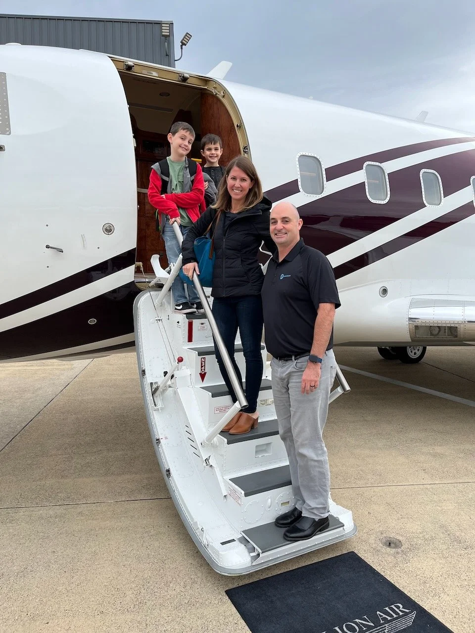 A group of people, including two children and two adults, standing on the steps leading to a private jet. The jet is parked on a runway, with one adult at the bottom of the stairs and the others above. The aircraft has a white exterior with maroon and gold stripes.