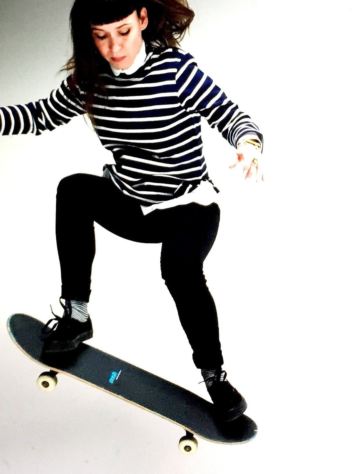 A woman with dark hair and bangs wearing a black and white striped shirt, black pants, and sneakers, skateboarding on a plain white background.