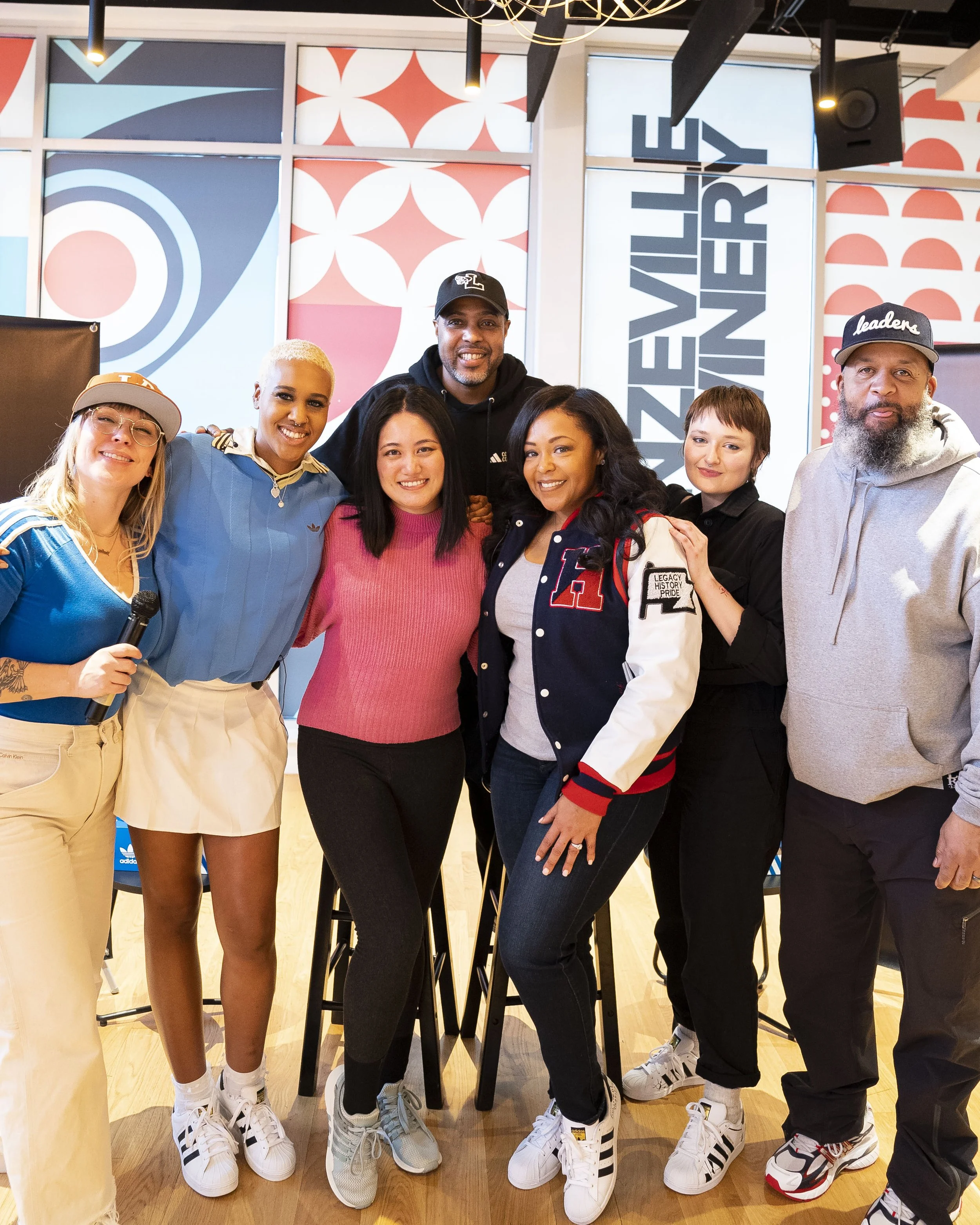A group of seven people standing together indoors with colorful patterned background, smiling at the camera, with a sign in the background reading 'WEU UNTIL WE WIN'. They are wearing casual clothing and Adidas sneakers.