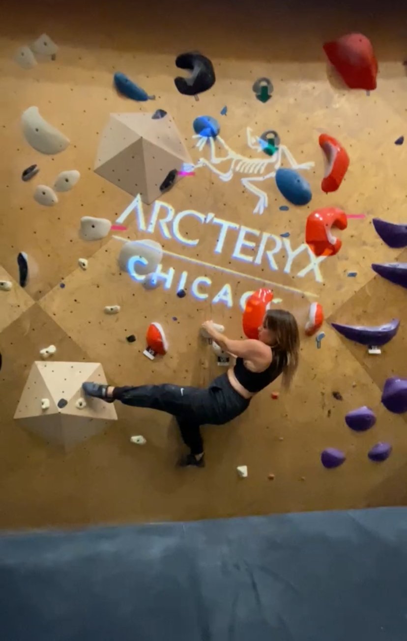 A person climbing an indoor rock wall at Arc'teryx Chicago, with multi-colored holds and a blue mat below.