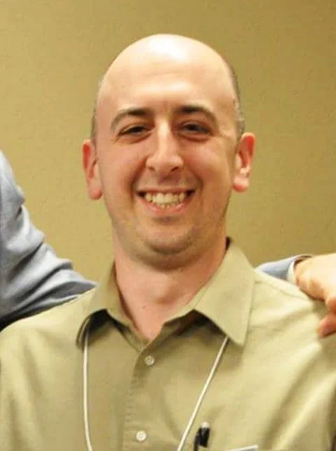 A smiling man with a bald head wearing a beige shirt and a lanyard, standing indoors.